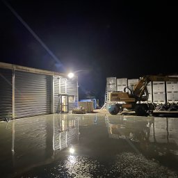 SEBASTIAN SÓJKA BASTEKPRO - Świeżo wylana, mokra posadzka przemysłowa przed budynkiem z metalowymi drzwiami rolowanymi, oświetlona reflektorem, z koparką Liebherr w tle i paletami z workami, wykonana nocą.