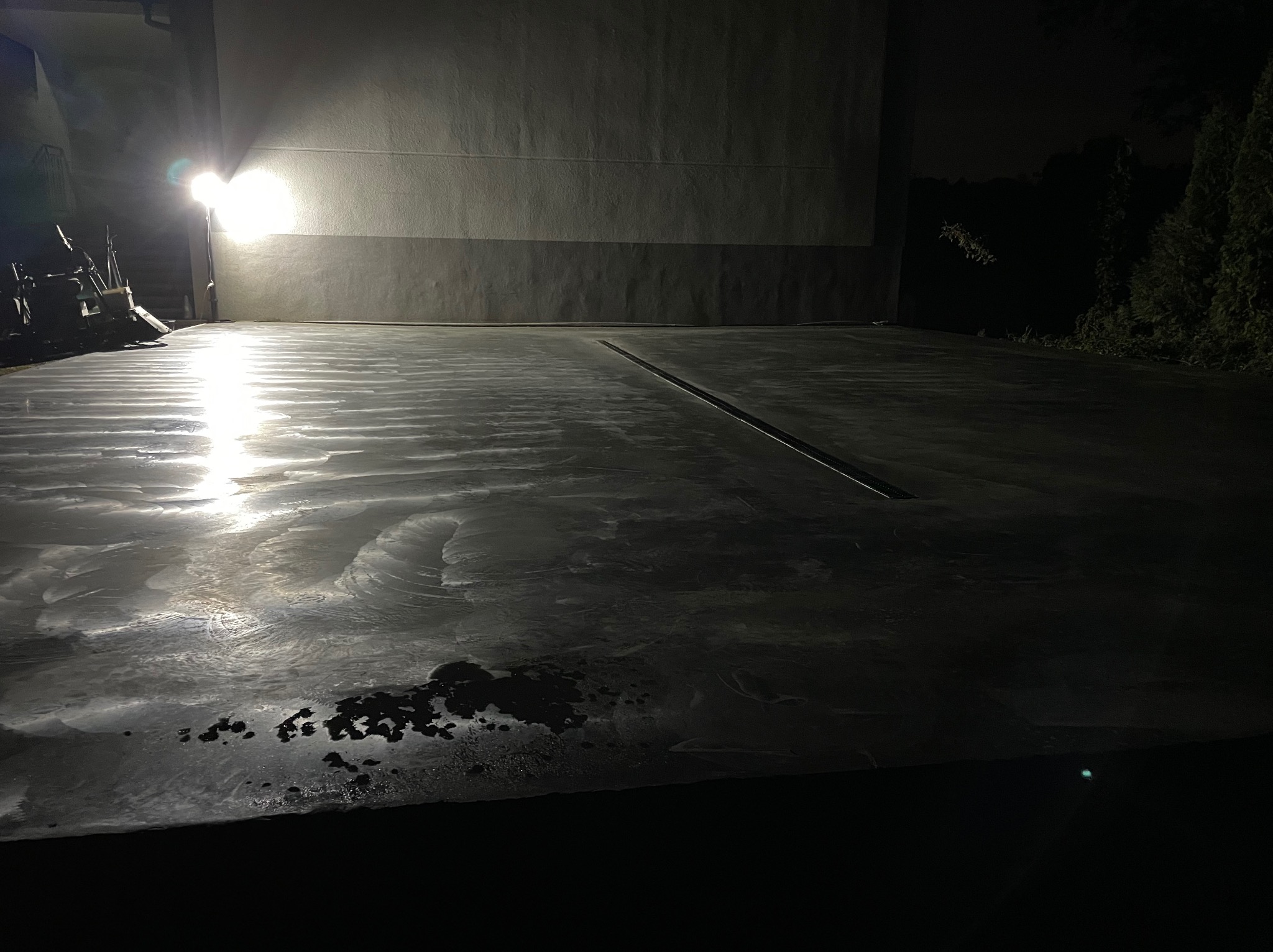 Wykończona, ciemna posadzka z widocznym odpływem liniowym, oświetlona punktowo, z ciemnymi plamami na powierzchni, na tle ściany i zieleni.