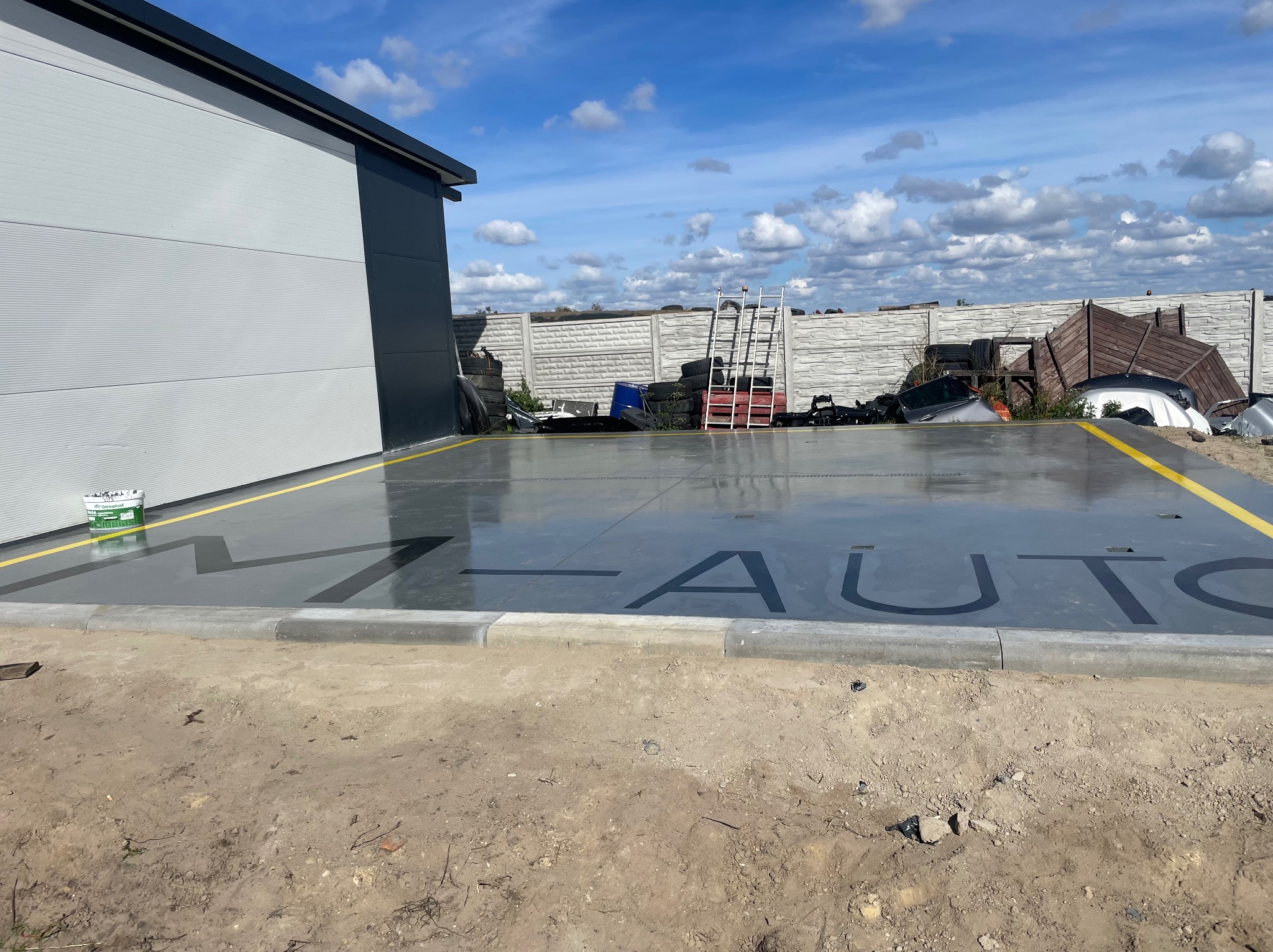 Szara posadzka z czarnym napisem 'AUTO' i żółtymi liniami, widok zewnętrzny z budynkiem w tle, na obrzeżach posadzki leżą opony i inne odpady.