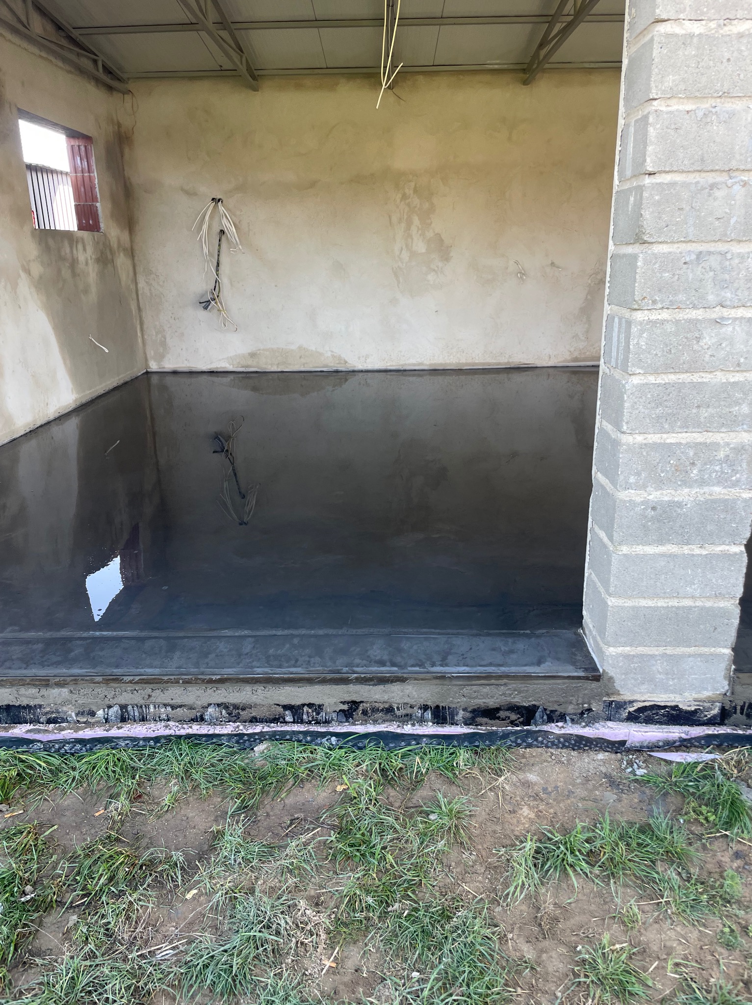 Świeżo wylana, mokra posadzka przemysłowa w surowym pomieszczeniu z widocznymi instalacjami elektrycznymi i fragmentami muru z bloczków betonowych, z zewnątrz widoczna trawa.