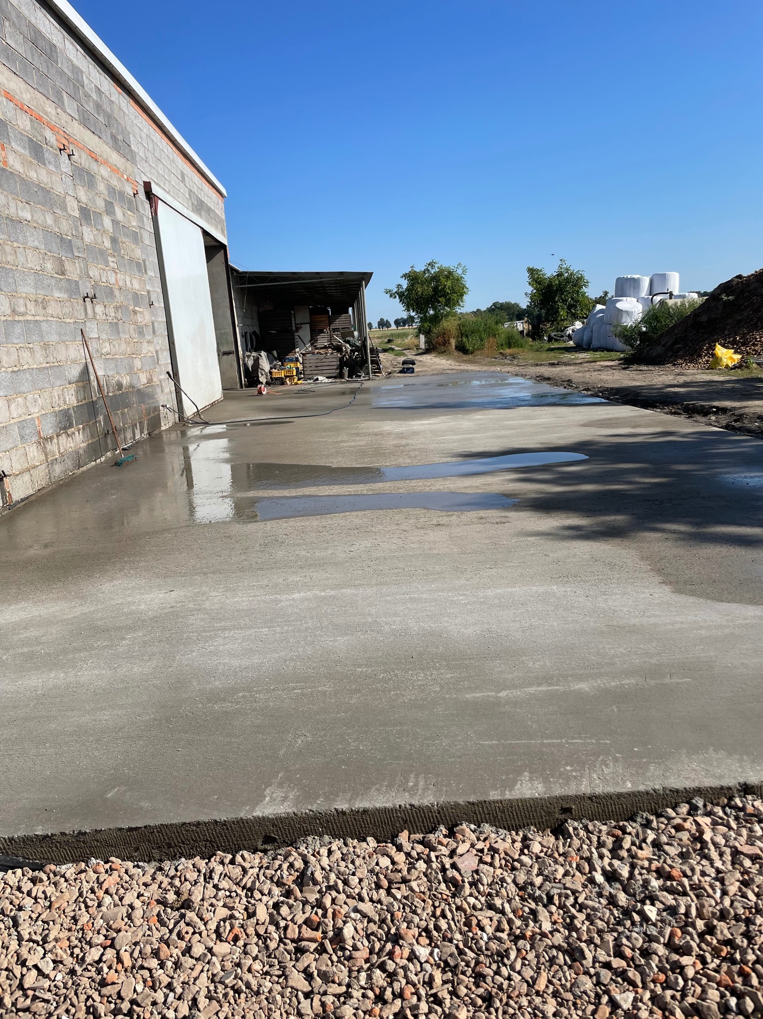 Świeżo wylana, gładka, betonowa powierzchnia z widocznymi śladami wody, przylegająca do ściany z szarej cegły i obszaru z drobnym żwirem, w tle budynek gospodarczy i błękitne niebo.