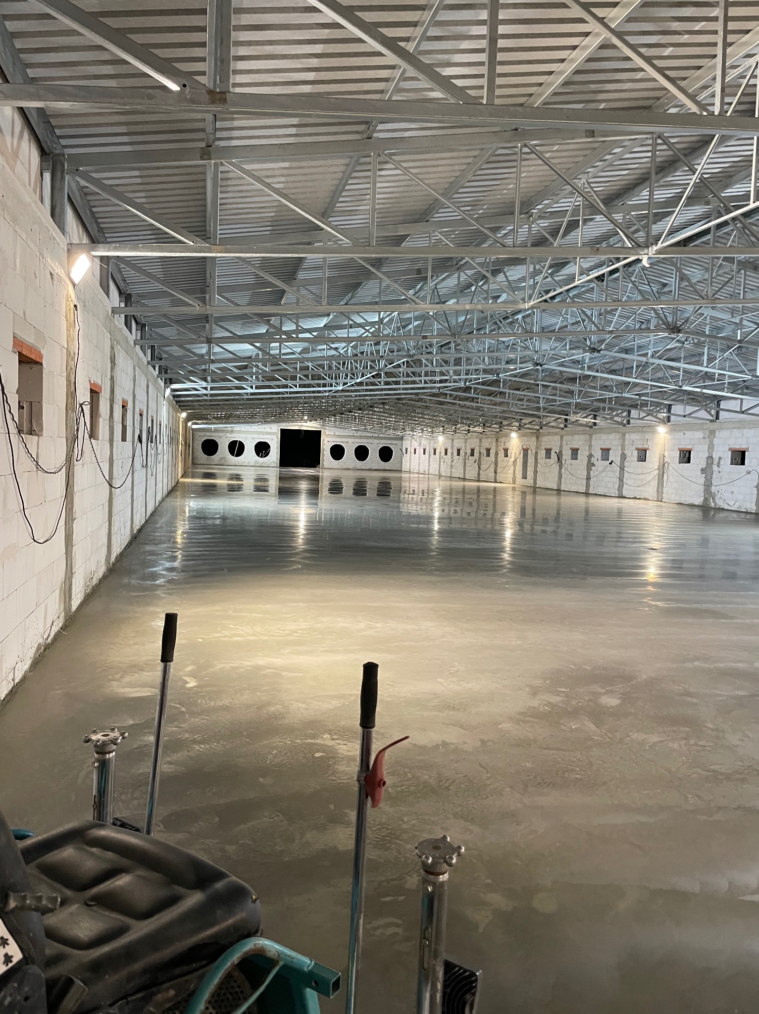Wnętrze hali produkcyjnej z wylewką posadzki przemysłowej w trakcie prac wykończeniowych, widoczna maszyna do zacierania betonu i ściany z pustaków betonowych.