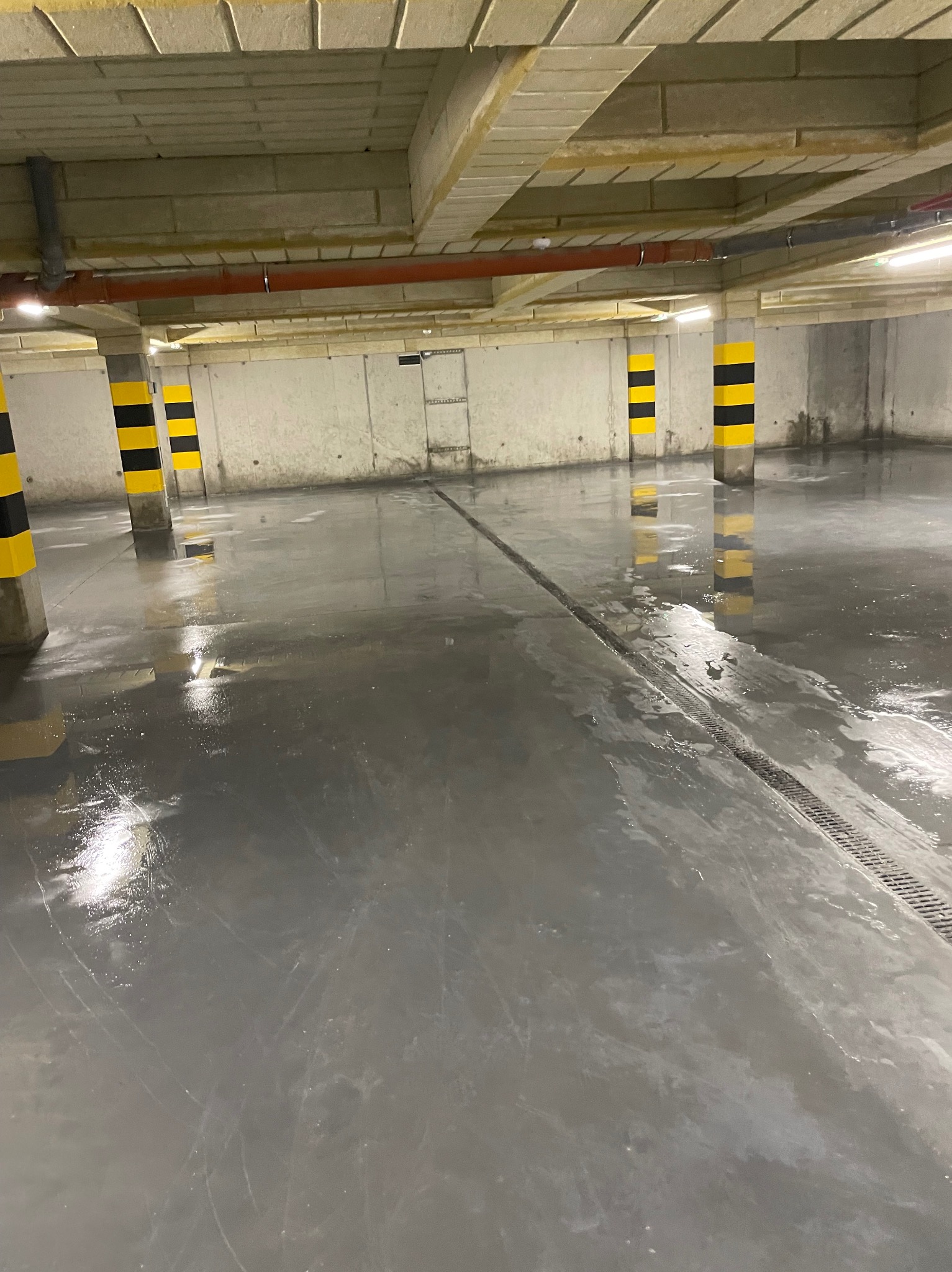 Świeżo wylana, mokra, szara podłoga w garażu podziemnym z żółto-czarnymi pasami na filarach i odpływem liniowym.