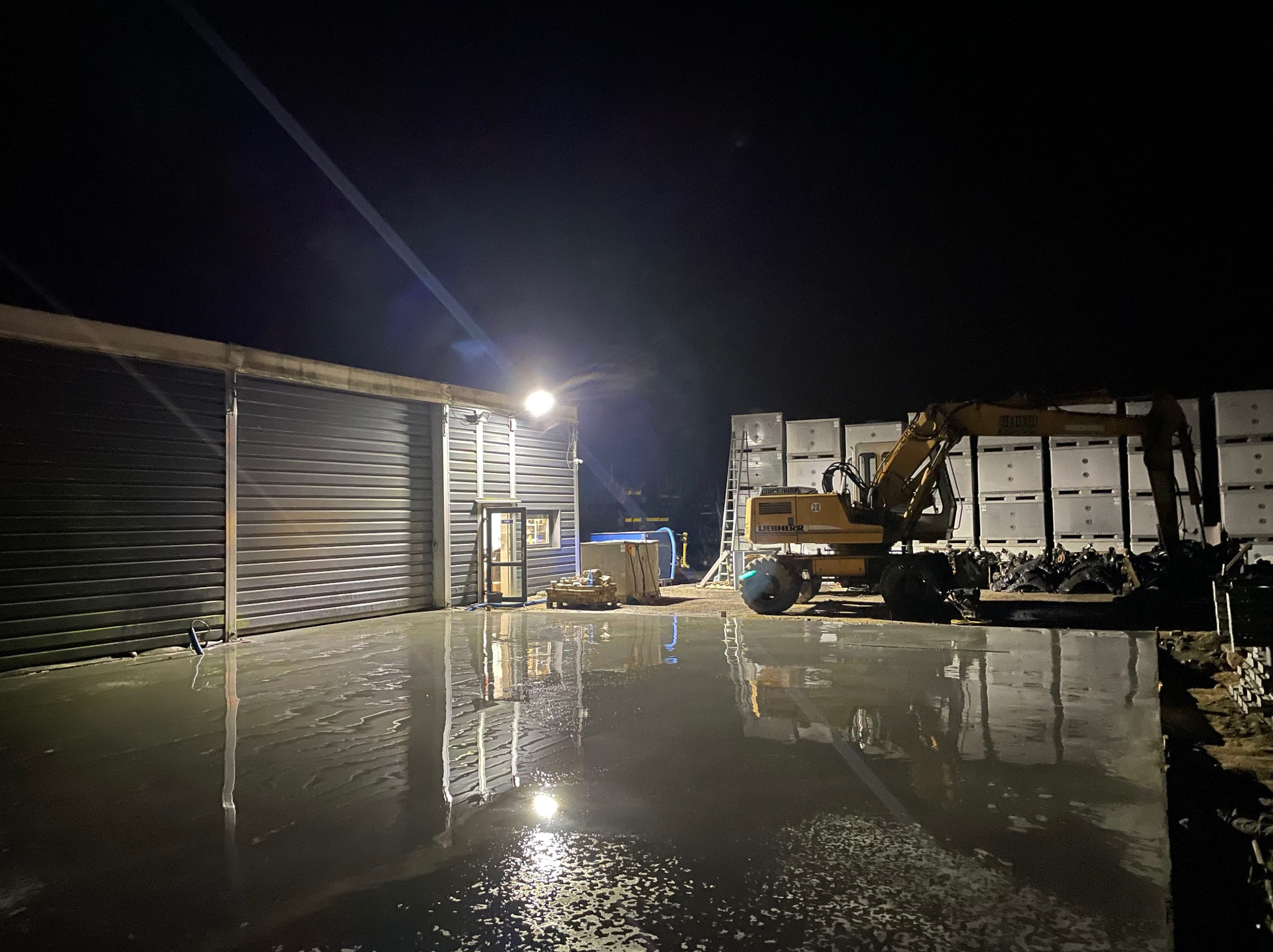 Świeżo wylana, mokra posadzka przemysłowa przed budynkiem z metalowymi drzwiami rolowanymi, oświetlona reflektorem, z koparką Liebherr w tle i paletami z workami, wykonana nocą.