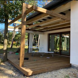 STA Design - Konstrukcja z belek zadaszenia tarasu drewnianego w trakcie budowy, widoczne narzędzia i materiały budowlane, surowy teren wokół.