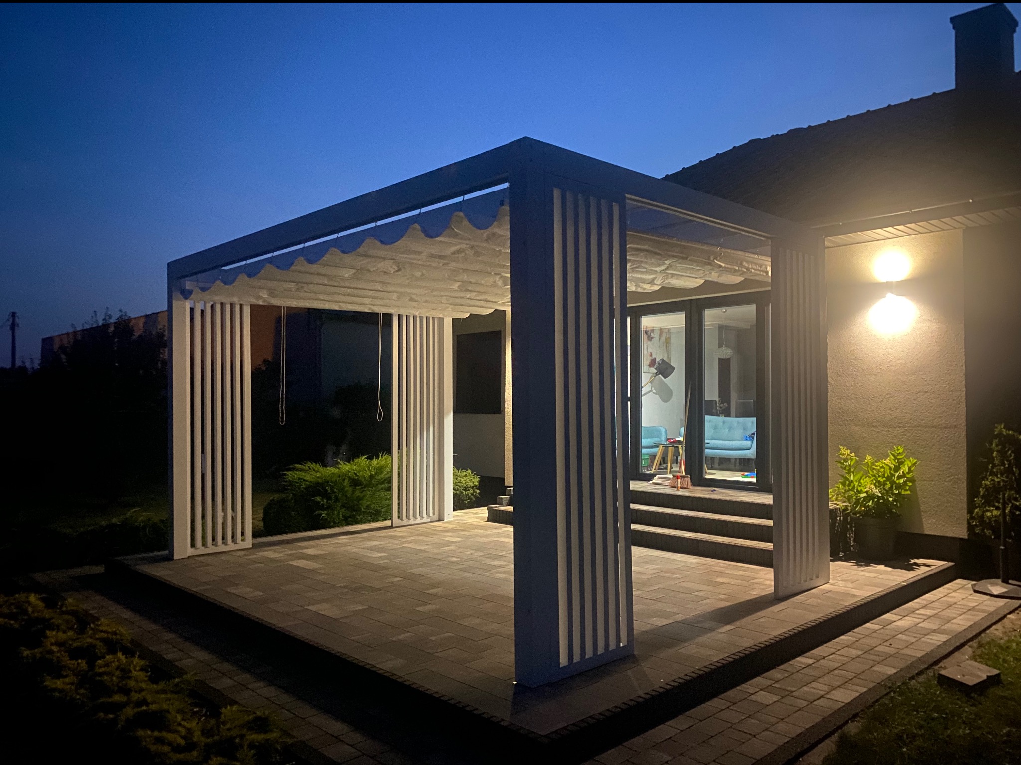 Nowoczesna, biała konstrukcja z zadaszeniem tekstylnym na tarasie z kostki brukowej, oświetlona ciepłym światłem, widok wieczorny.