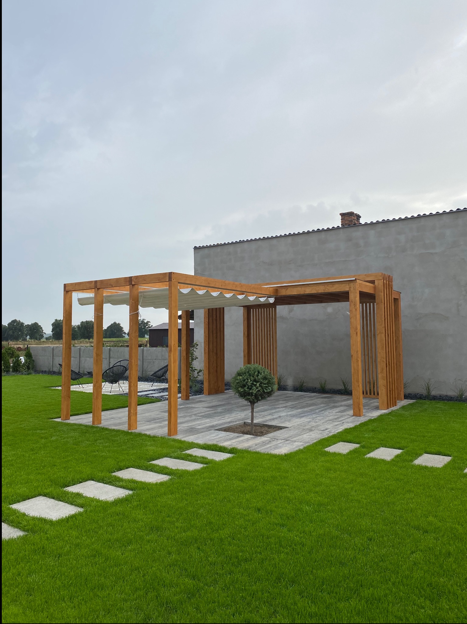 Drewniana pergola z kremową, falowaną markizą na betonowej podłodze z małym drzewkiem pośrodku, widok z trawnika z kamienną ścieżką, w tle szary budynek.