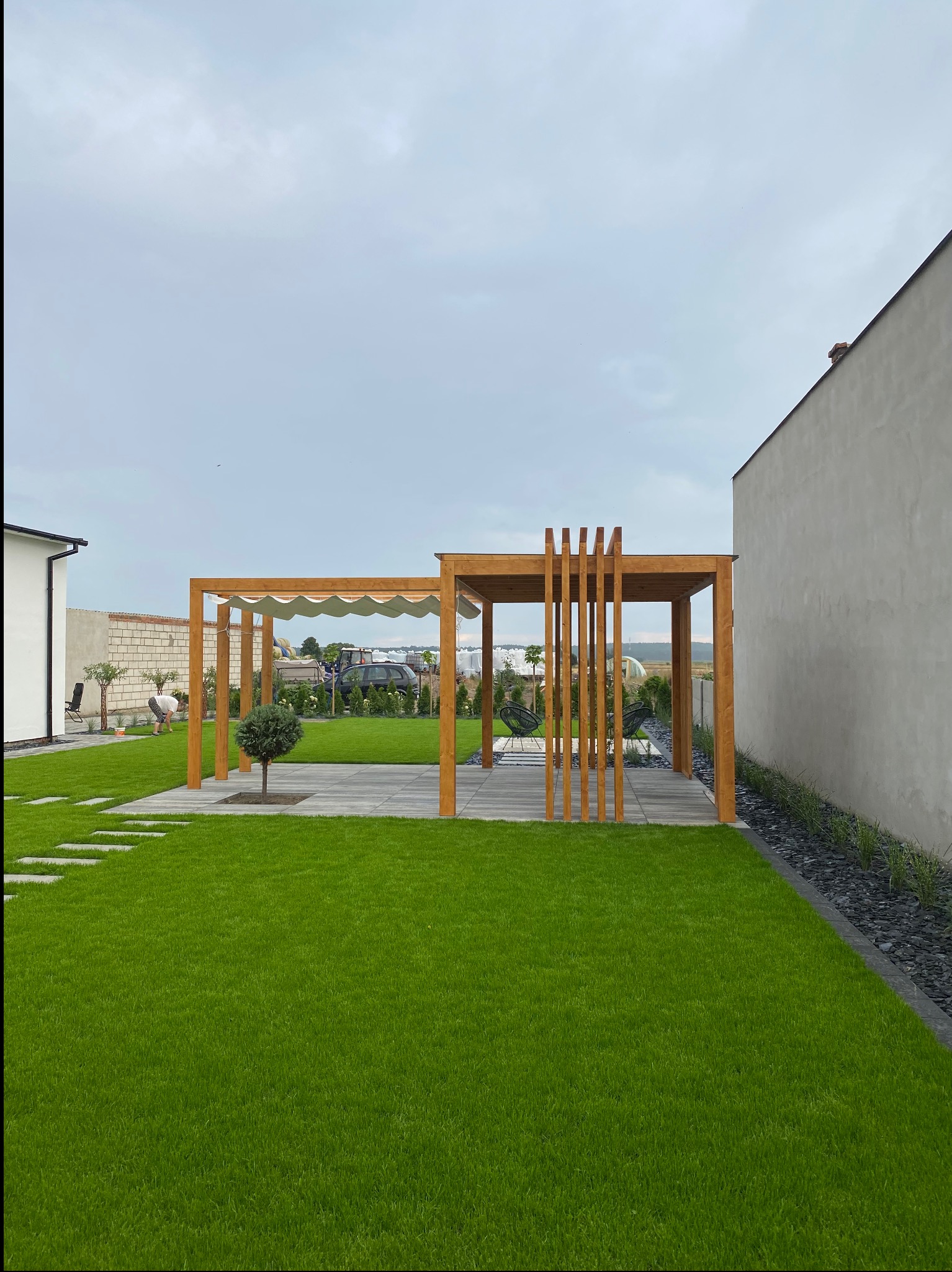 Dwie drewniane konstrukcje ogrodowe z elementami zacieniającymi, połączone ścieżką z płyt betonowych na trawniku, w tle osoba pielęgnująca rośliny.