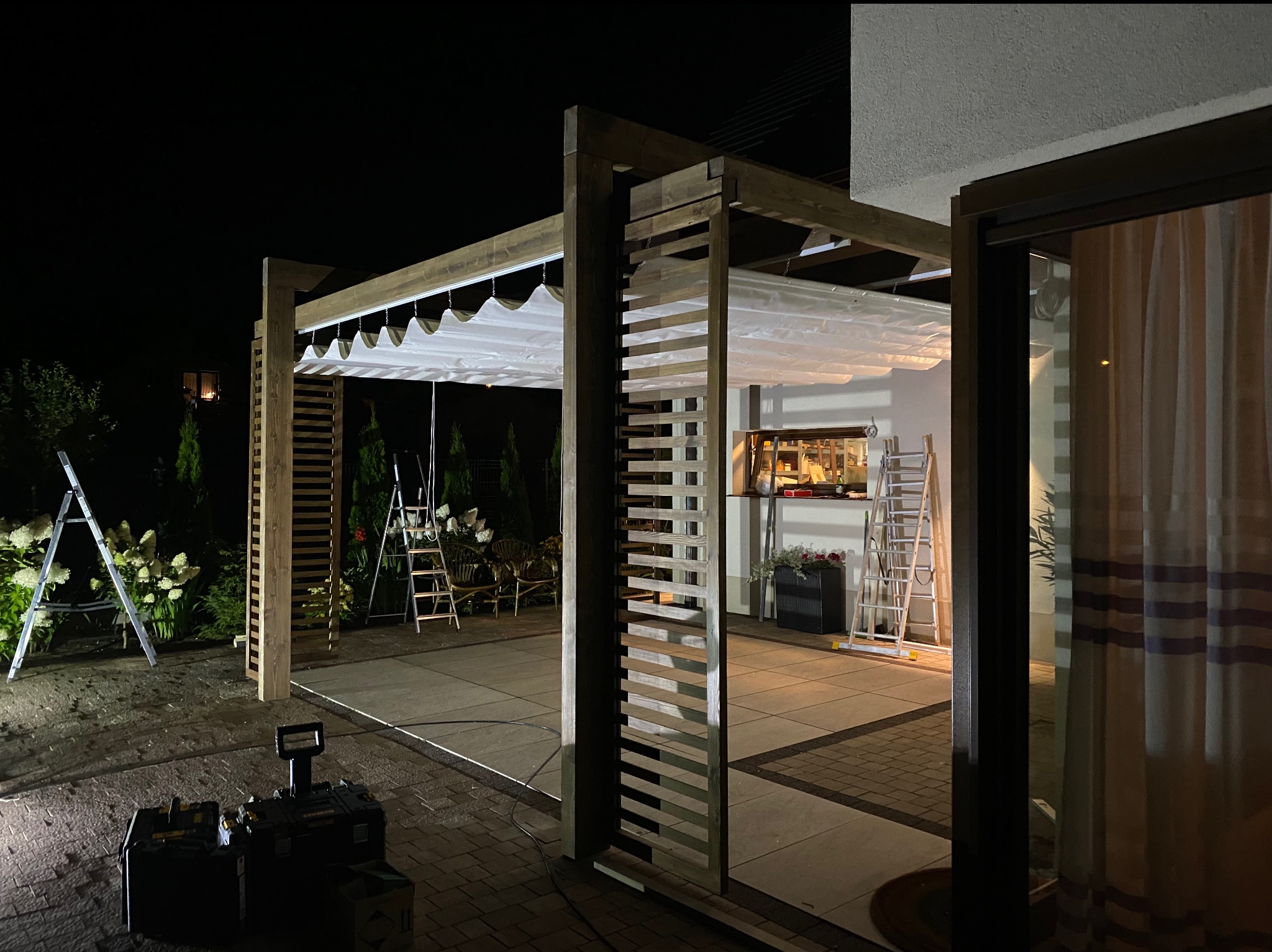 Drewniana konstrukcja z białym zadaszeniem w ogrodzie nocą, widoczne narzędzia i drabiny, oświetlenie robocze.