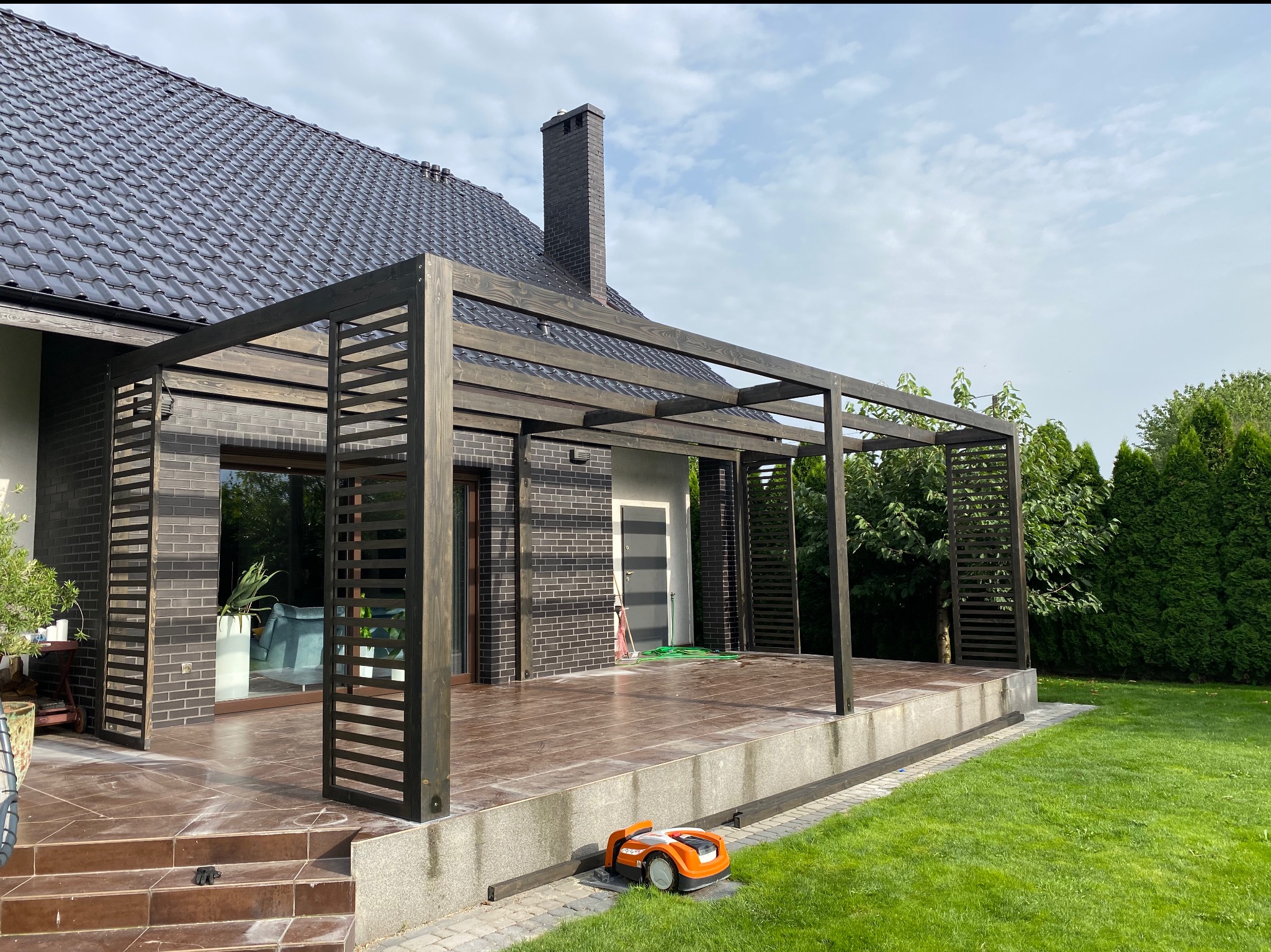 Nowoczesna, ciemna konstrukcja z drewna z ażurowymi ścianami, przylegająca do domu z ciemnej cegły, z robotem koszącym na trawniku.