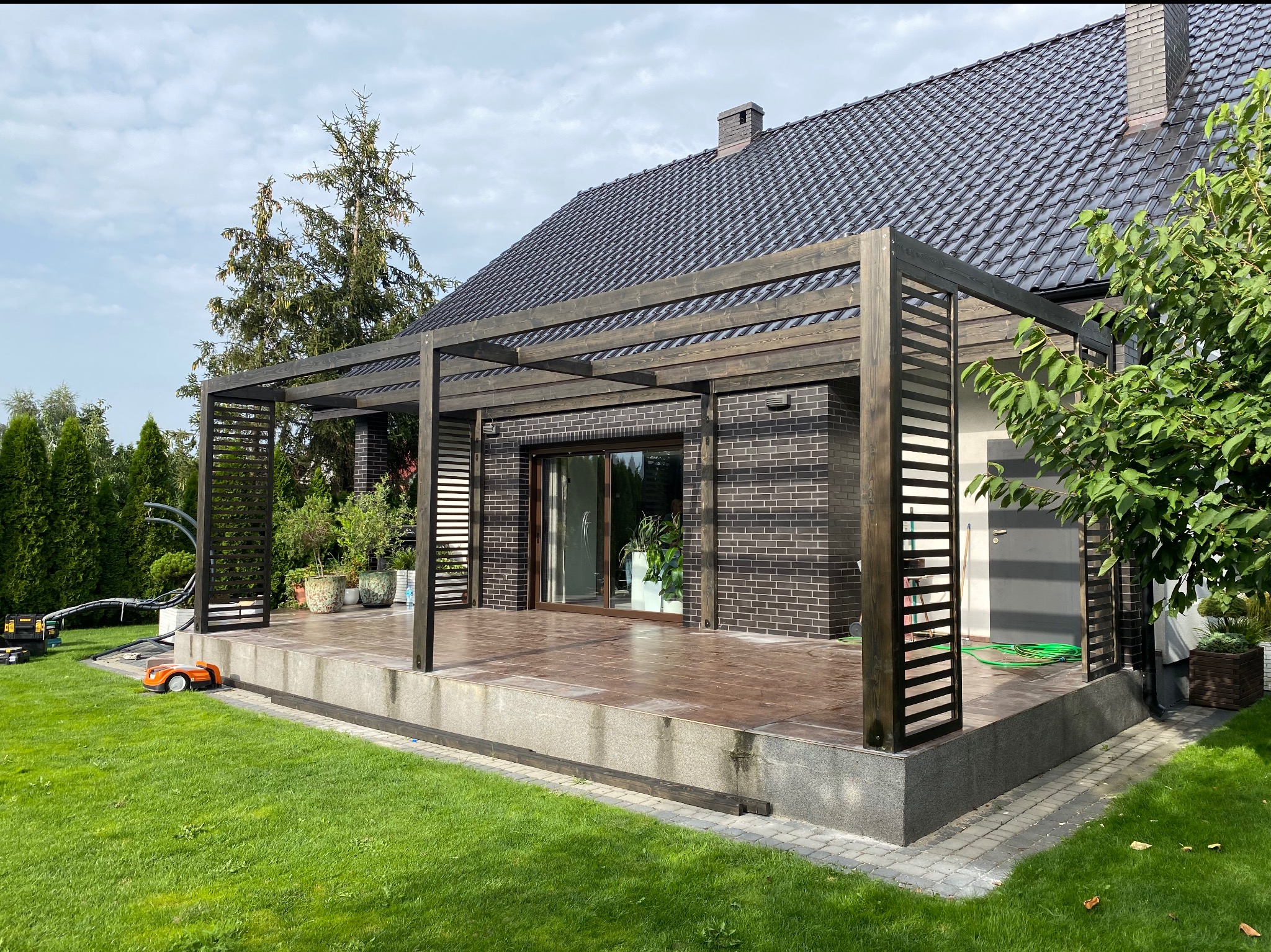Nowoczesna konstrukcja z ciemnego drewna, tworząca zadaszenie tarasu przy domu z ciemnej cegły, widoczny robot koszący na trawniku.