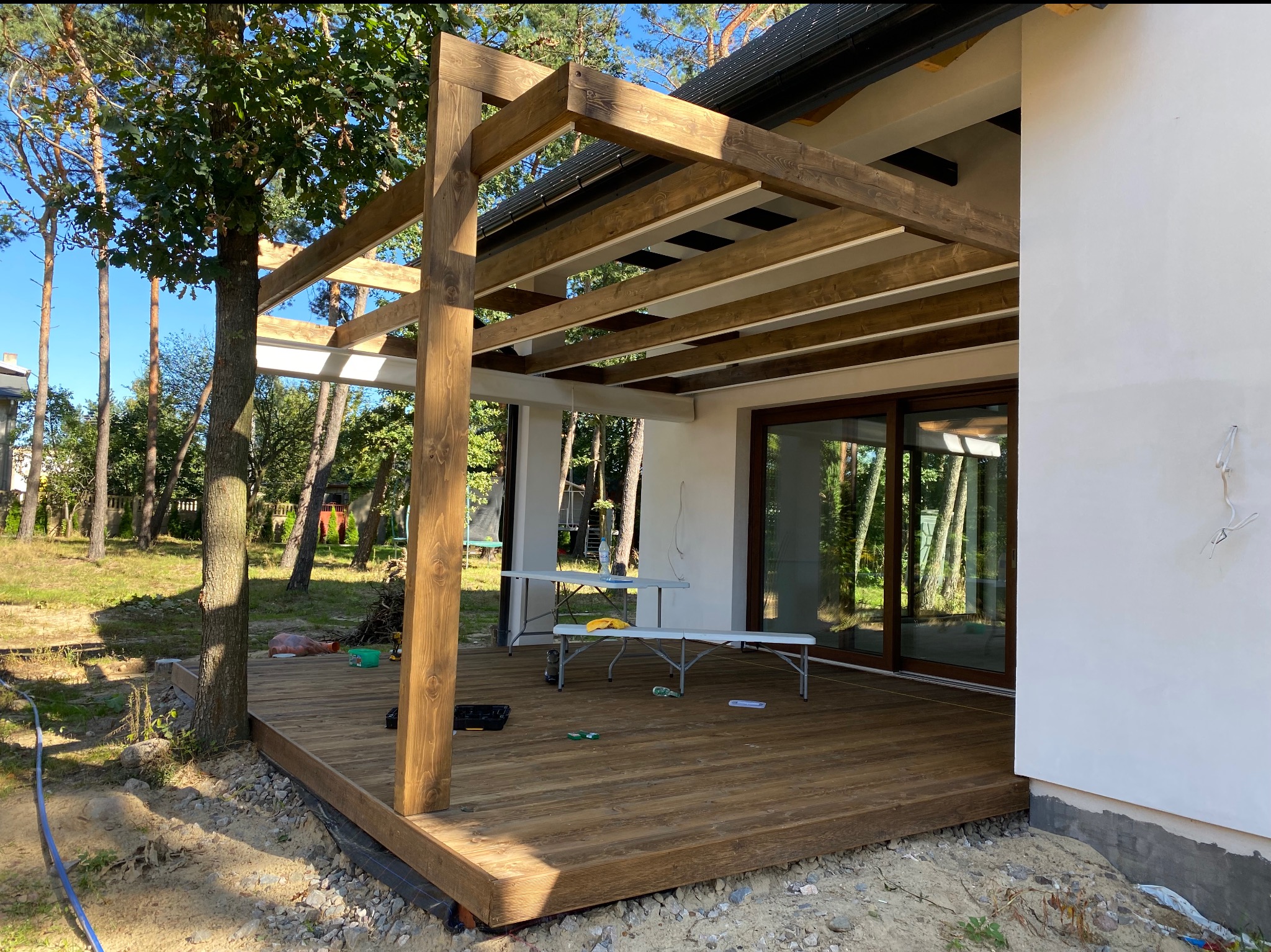 Konstrukcja z belek zadaszenia tarasu drewnianego w trakcie budowy, widoczne narzędzia i materiały budowlane, surowy teren wokół.