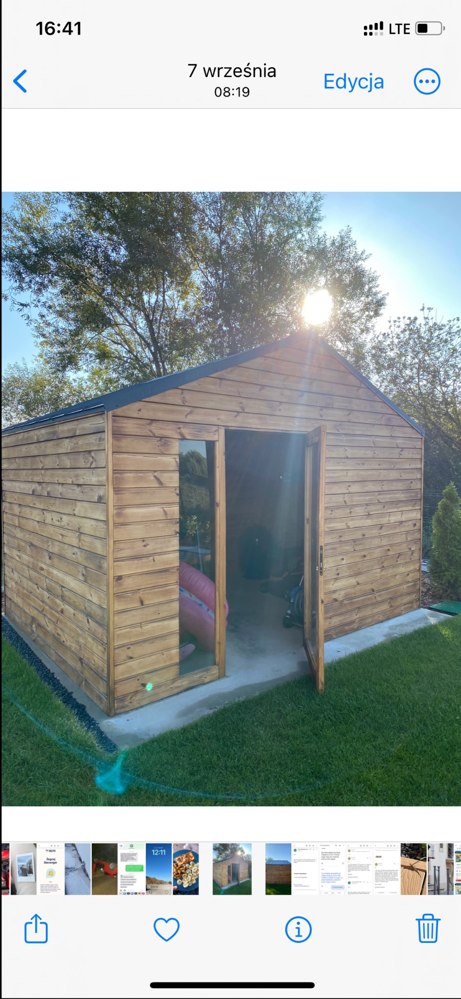 Drewniany domek narzędziowy z otwartymi drzwiami, widok na wnętrze z dmuchanym materacem i rowerem, trawnik i drzewa w tle, słoneczny dzień.