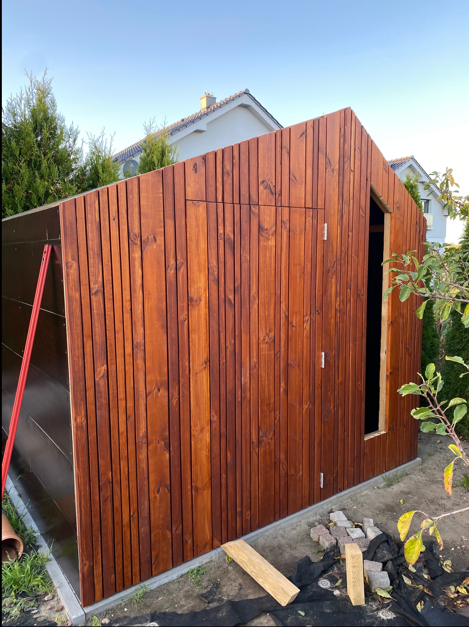 Brązowa drewniana konstrukcja z pionowych desek w trakcie budowy, widoczne drzwi i okno bez wypełnienia, cegły i drewniane belki leżą na ziemi.