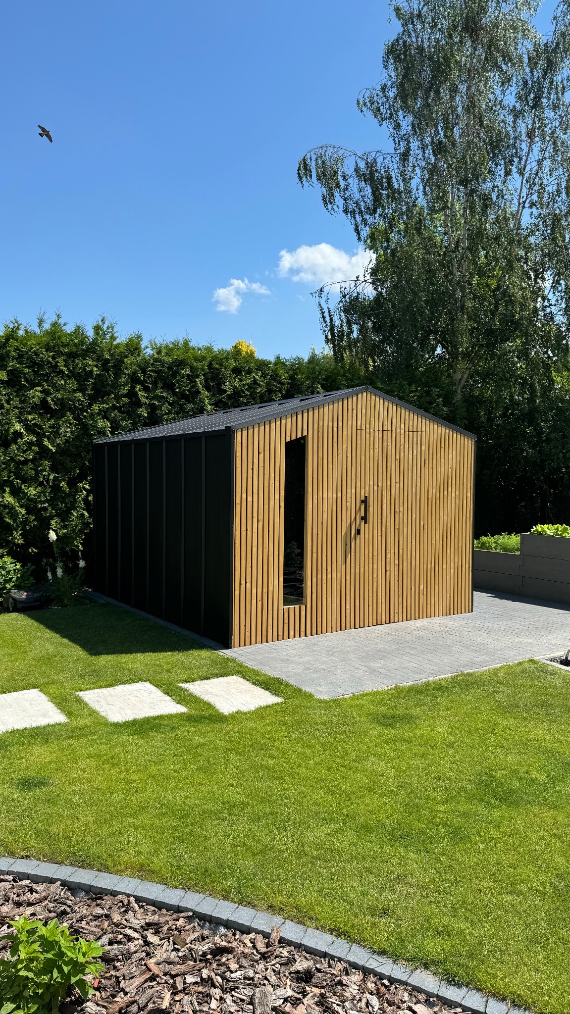Nowoczesna konstrukcja z drewna i czarnych paneli, z pionowymi deskami i płaskim dachem, na tle zielonego trawnika i żywopłotu, w słoneczny dzień.
