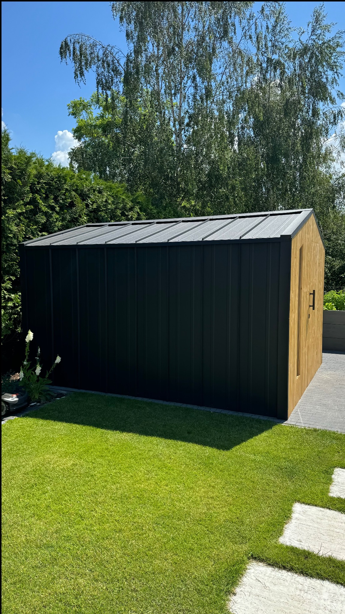 Nowoczesna, prostokątna konstrukcja z czarnymi ścianami i drewnianą elewacją, zlokalizowana w ogrodzie z zielonym trawnikiem i drzewami w tle.