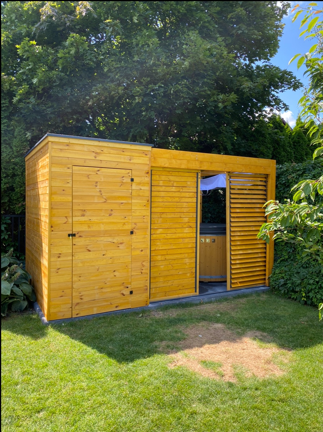 Nowoczesna, żółta konstrukcja z drewna, składająca się z zamykanego pomieszczenia z drzwiami i otwartej części z żaluzjami, pod którą widać drewnianą wannę z hydromasażem, otoczona zielenią ogrodu.