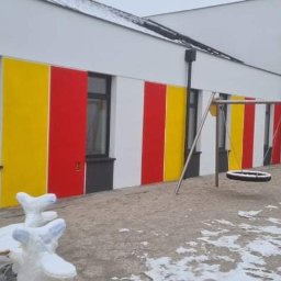 Marcin Zarzycki - Budynek z kolorowymi pasami (żółty, czerwony, biały) na elewacji, plac zabaw z huśtawkami. Częściowo ośnieżony teren. Widoczne okna i lampa na ścianie.