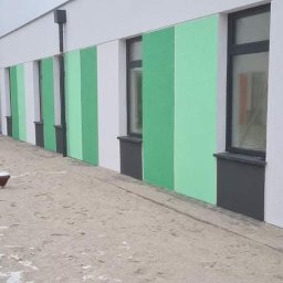 Marcin Zarzycki - Ściana budynku pomalowana w pasy biało-zielone, z oknami w ciemnych ramach. Na pierwszym planie plac zabaw pokryty piaskiem i śniegiem.