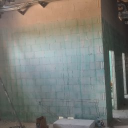 Marcin Zarzycki - Wnętrze w trakcie budowy z wymurowanymi ścianami z bloczków. Widoczny pracownik na drabinie montujący instalacje pod sufitem.