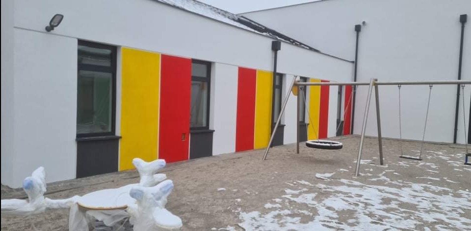 Budynek z kolorowymi pasami (żółty, czerwony, biały) na elewacji, plac zabaw z huśtawkami. Częściowo ośnieżony teren. Widoczne okna i lampa na ścianie.