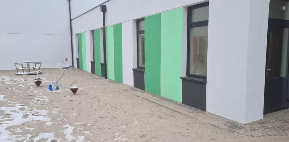 Ściana budynku pomalowana w pasy biało-zielone, z oknami w ciemnych ramach. Na pierwszym planie plac zabaw pokryty piaskiem i śniegiem.