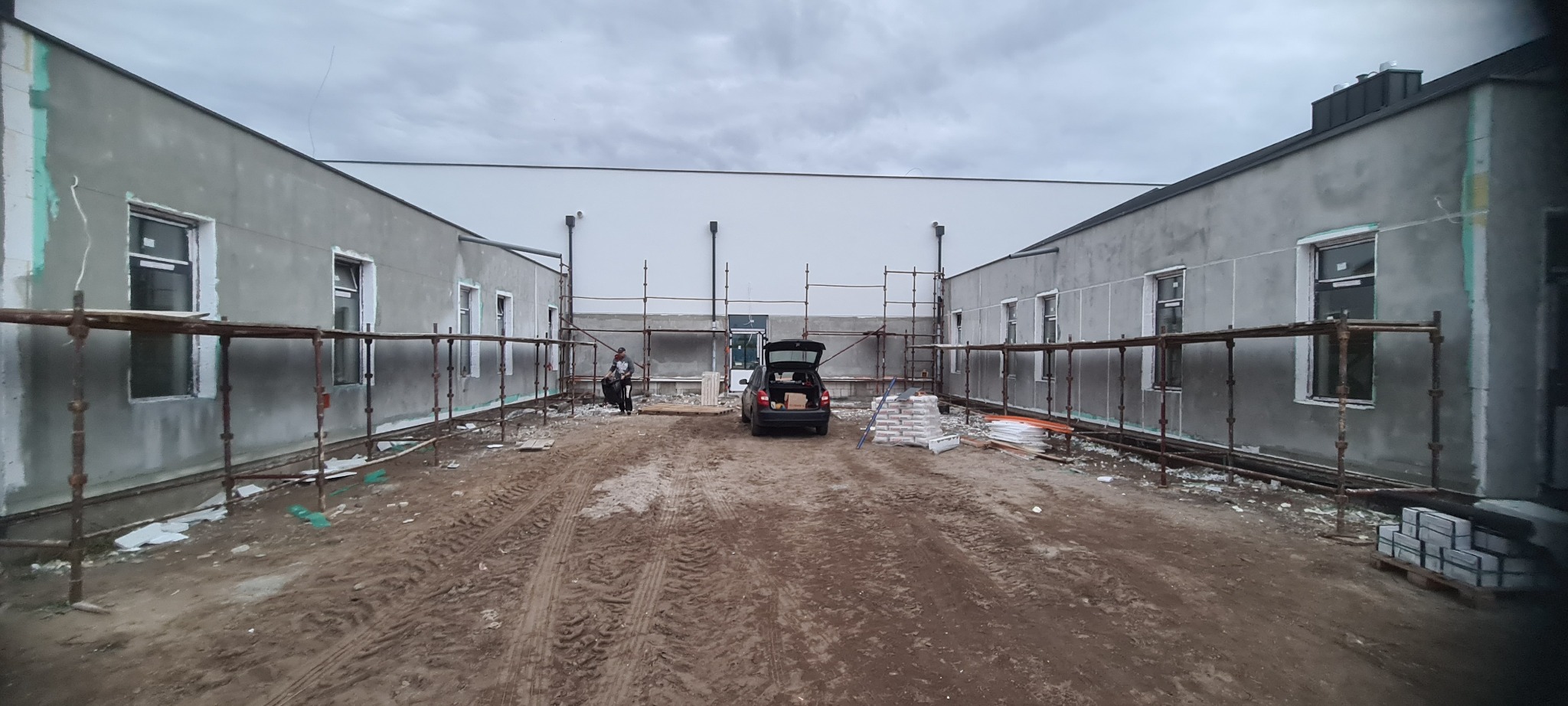 Budynek w trakcie prac murarskich: ściany otynkowane, rusztowania, materiały budowlane. Widoczny pracownik i samochód z otwartym bagażnikiem. Pochmurne niebo.