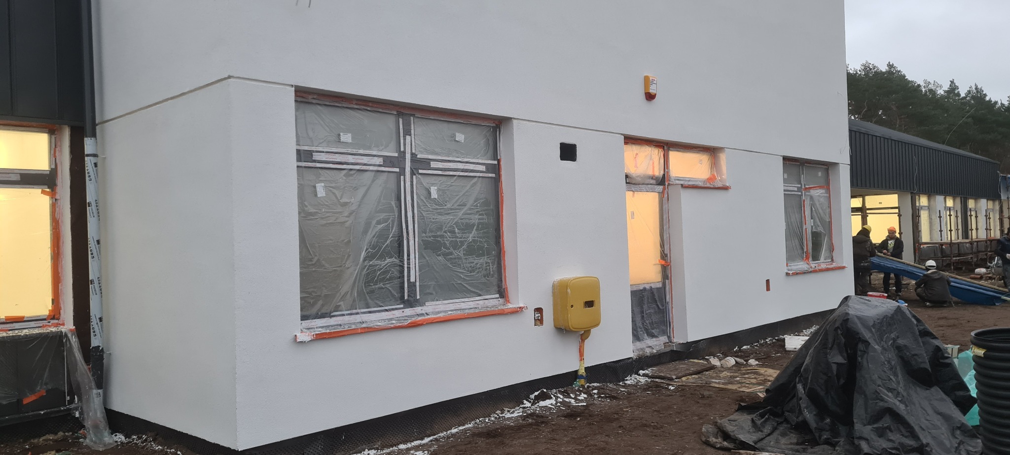 Biała elewacja budynku w trakcie wykończenia, okna zabezpieczone folią, widoczni pracownicy budowlani, teren budowy z elementami instalacji.