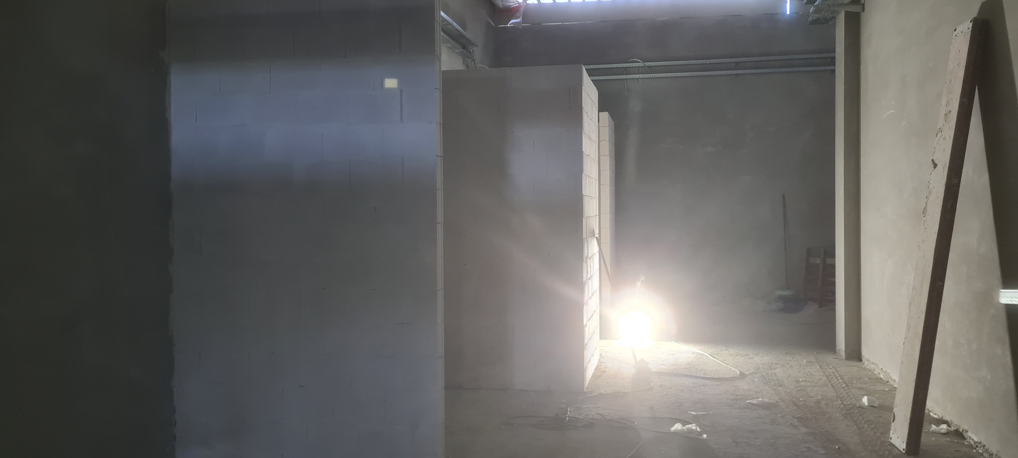 Wnętrze w trakcie budowy: wzniesione ściany z betonu komórkowego, widoczne instalacje pod sufitem i prowizoryczne oświetlenie. Surowy stan, przygotowanie do dalszych prac.