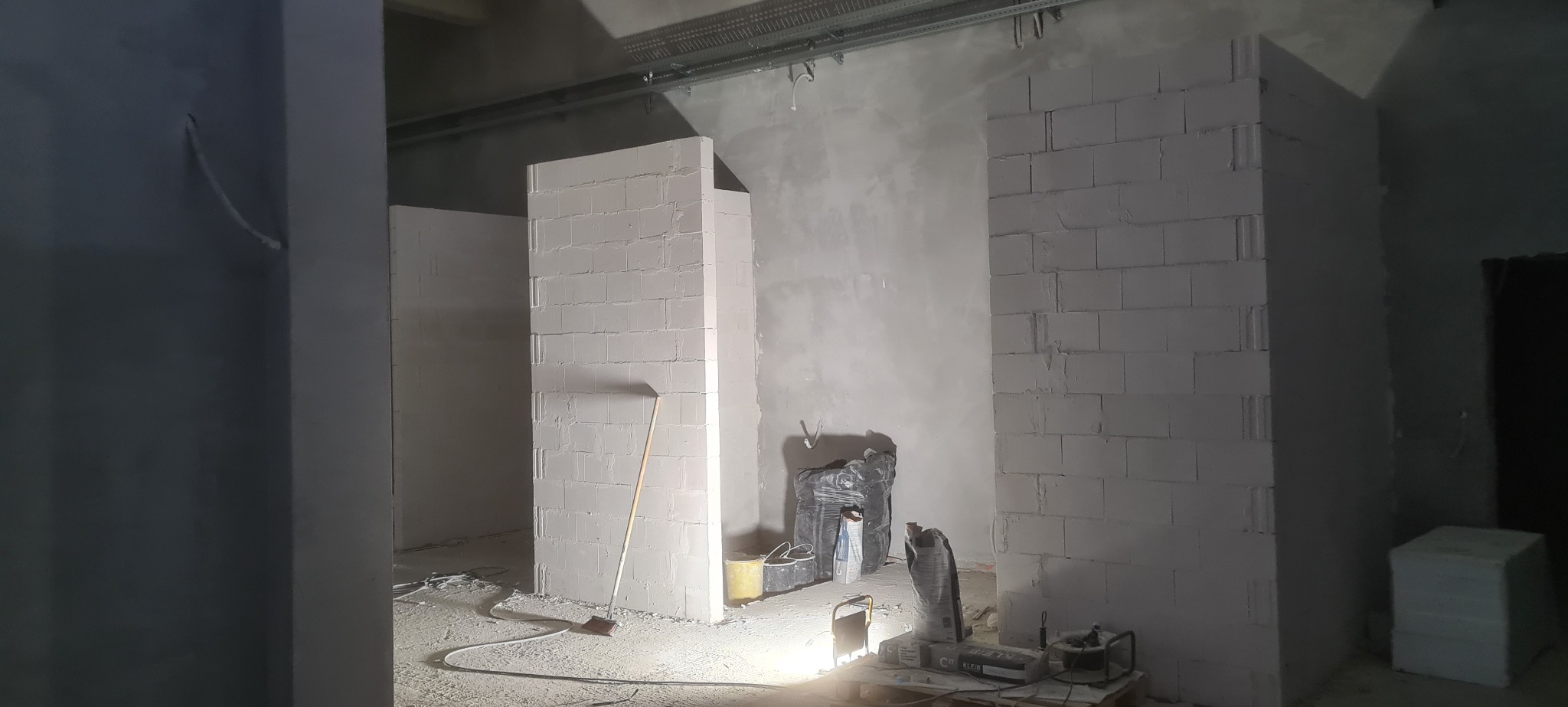 Wnętrze w trakcie budowy: ściany z białych bloczków, narzędzia i materiały budowlane, surowy betonowy strop z instalacjami. Widoczny etap murowania ścian działowych.