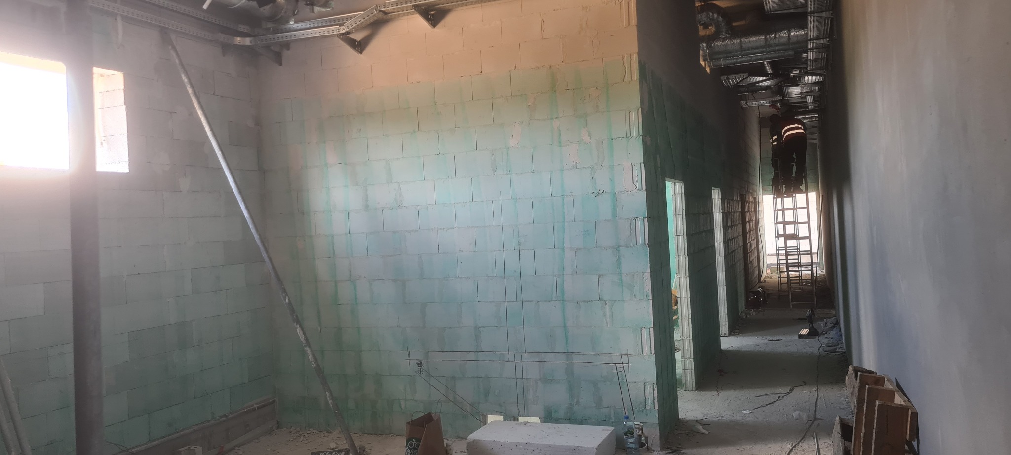 Wnętrze w trakcie budowy z wymurowanymi ścianami z bloczków. Widoczny pracownik na drabinie montujący instalacje pod sufitem.