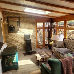 MACIEJCZEK EUGENIUSZ ZAKŁAD STOLARSKI-EXPORT-IMPORT Maciejczek Eugeniusz - Wnętrze przeszklonej altany z ceglaną ścianą, piecem typu koza, drewnianym stropem i meblami w stylu vintage, widok na ogród z ułożonym drewnem.