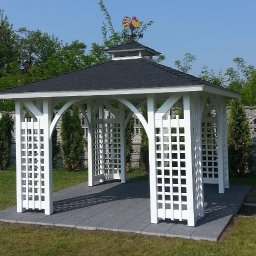 MACIEJCZEK EUGENIUSZ ZAKŁAD STOLARSKI-EXPORT-IMPORT Maciejczek Eugeniusz - Biała altana ogrodowa z czarnym dachem, zwieńczona wiatrowskazem z kogutem, na szarym podłożu z kostki brukowej, otoczona zielenią.