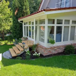 Zadaszony taras z drewnianymi schodami, ceglaną podstawą i białymi ramami okiennymi, otoczony zielenią i lampami ogrodowymi.