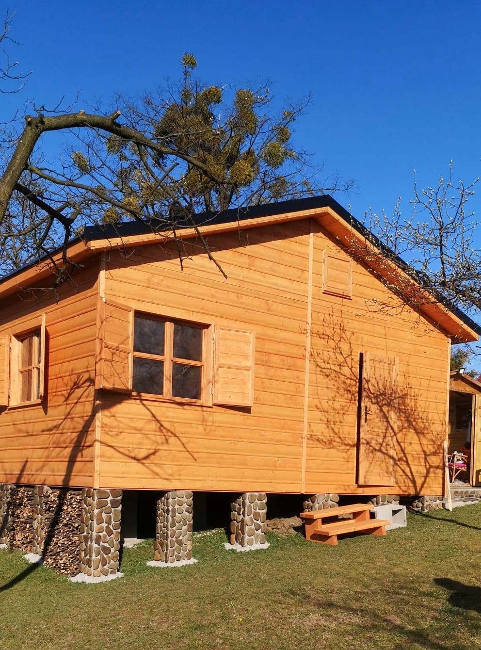 Drewniana altana na kamiennych fundamentach, z otwartym oknem i małymi schodkami wejściowymi, otoczona drzewami w słoneczny dzień.