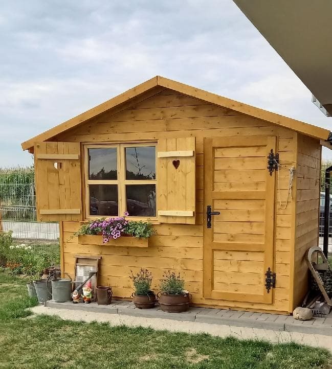 Mała, drewniana konstrukcja ogrodowa z oknem, okiennicami z wyciętymi sercami i skrzynką z fioletowymi kwiatami, otoczona dekoracjami ogrodowymi i roślinami w donicach.