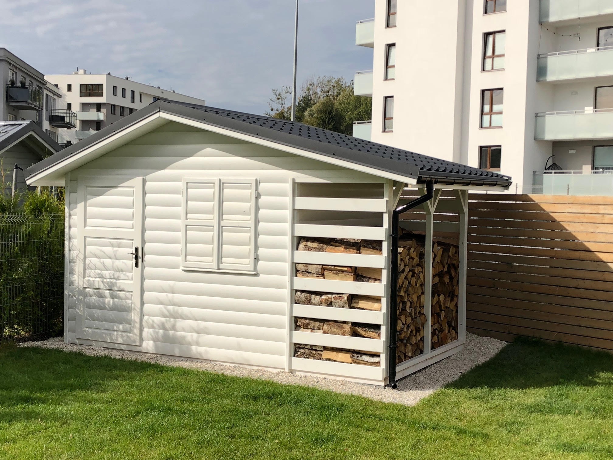 Biała altana ogrodowa z miejscem na składowanie drewna, widok na nowoczesne budynki mieszkalne w tle.