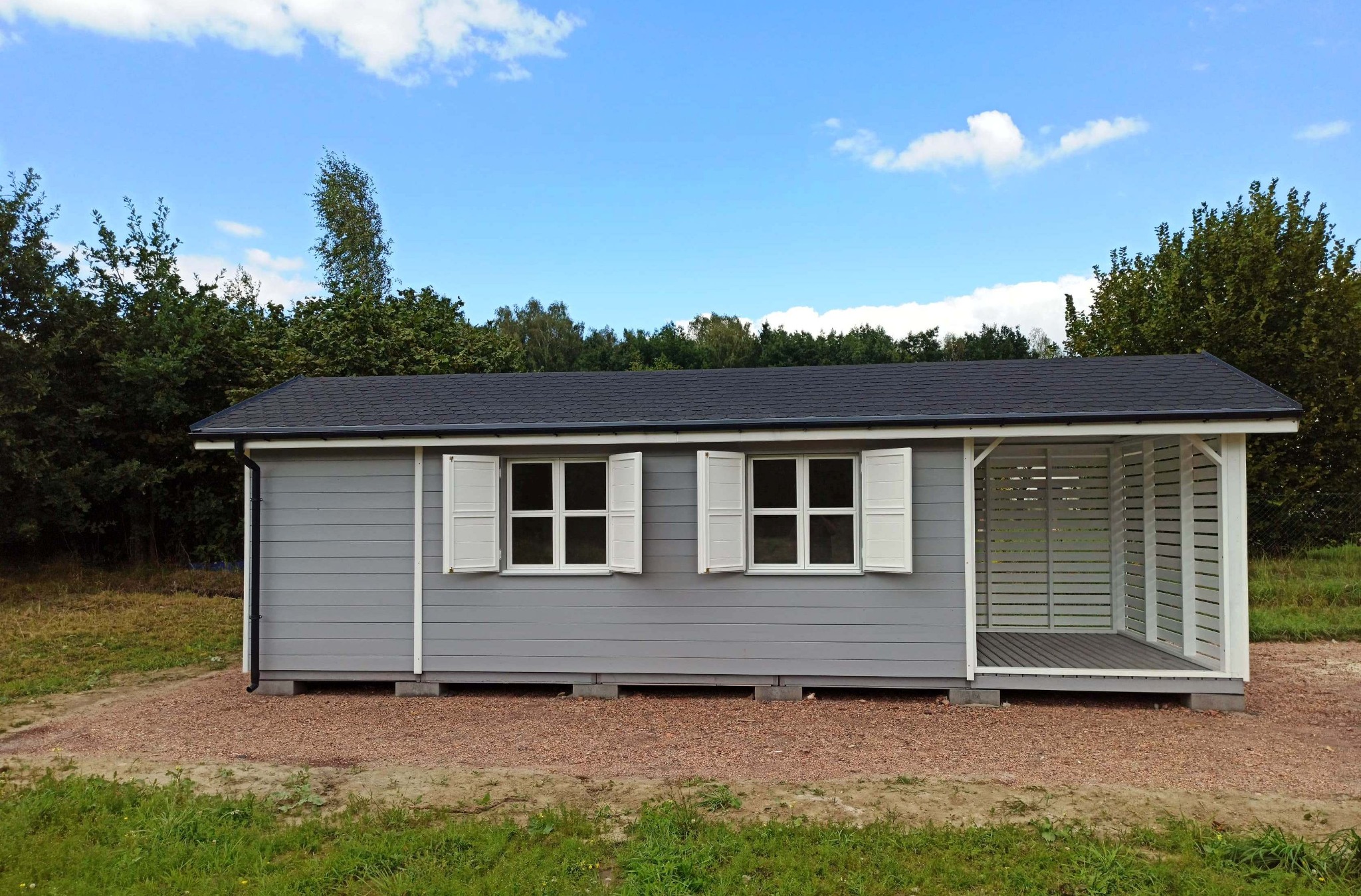 Szara konstrukcja z dwoma oknami z białymi okiennicami i otwartą przestrzenią z białymi, poziomymi listwami, otoczona zielenią i żwirem.