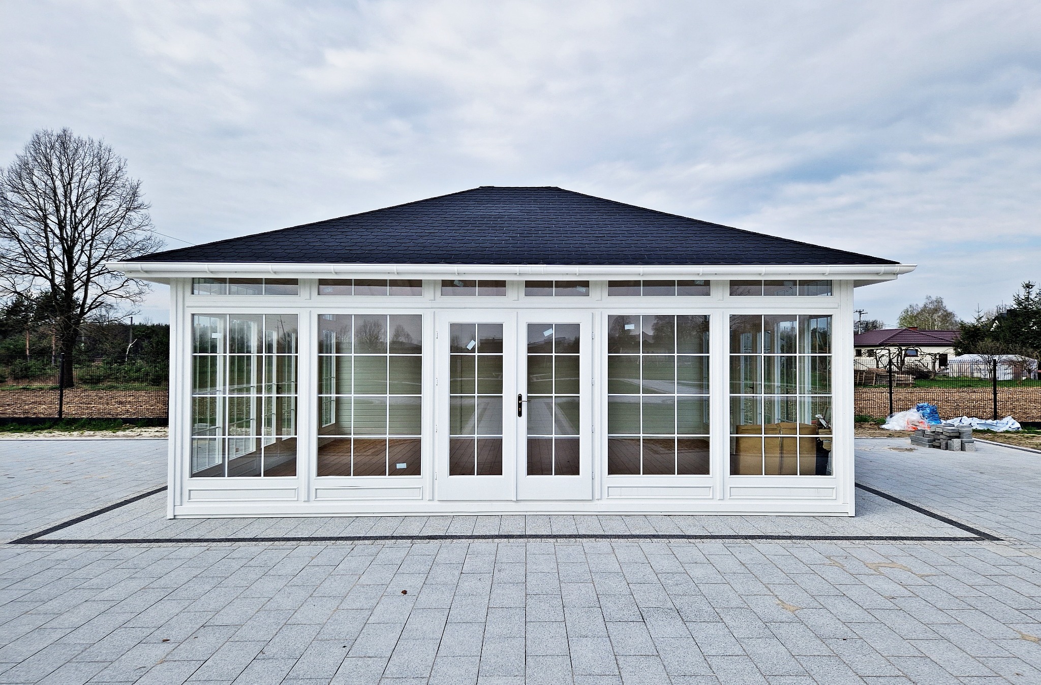 Elegancka, przeszklona altana ogrodowa z dwuspadowym, ciemnym dachem, otoczona szarą kostką brukową i zielenią w tle.