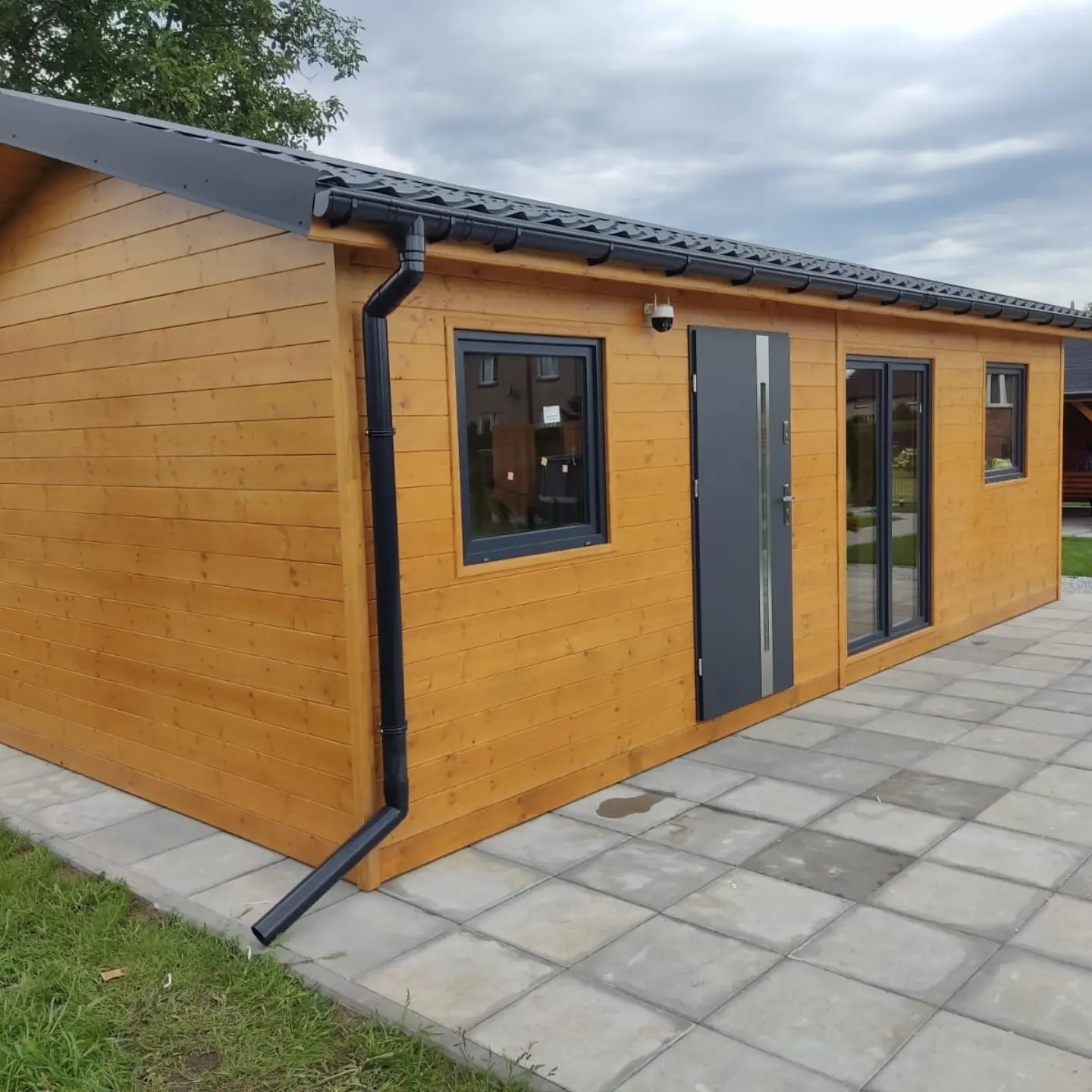 Nowoczesna, drewniana konstrukcja z szarymi elementami: drzwi, okna, dach i rynny. Budowla stoi na wyłożonym kostką brukową terenie.