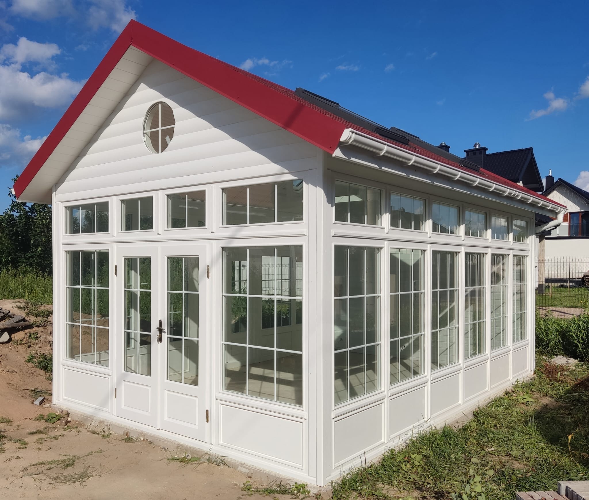 Biała konstrukcja z czerwonym dachem i licznymi oknami, przypominająca oranżerię, na tle błękitnego nieba.