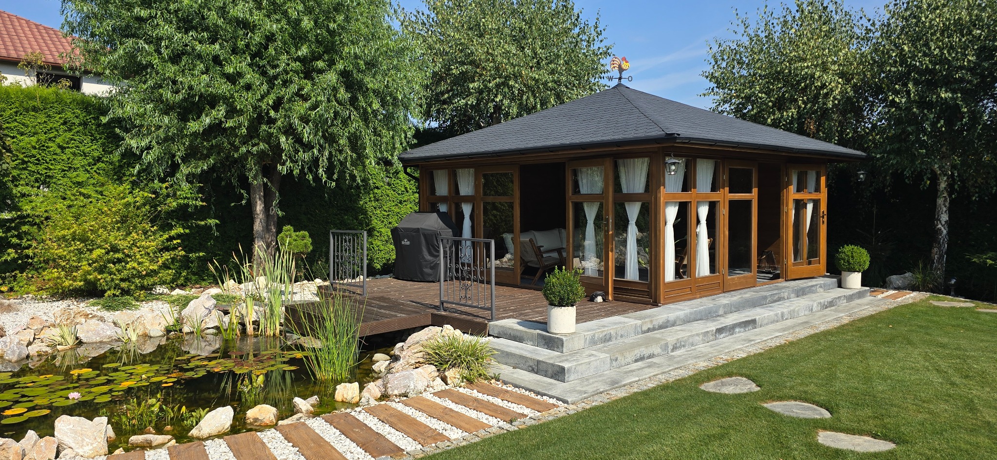 Ogród z drewnianą altaną z ciemnym dachem i wiatrowskazem, stawem z liliami wodnymi, drewnianym tarasem i grillem na zewnątrz.
