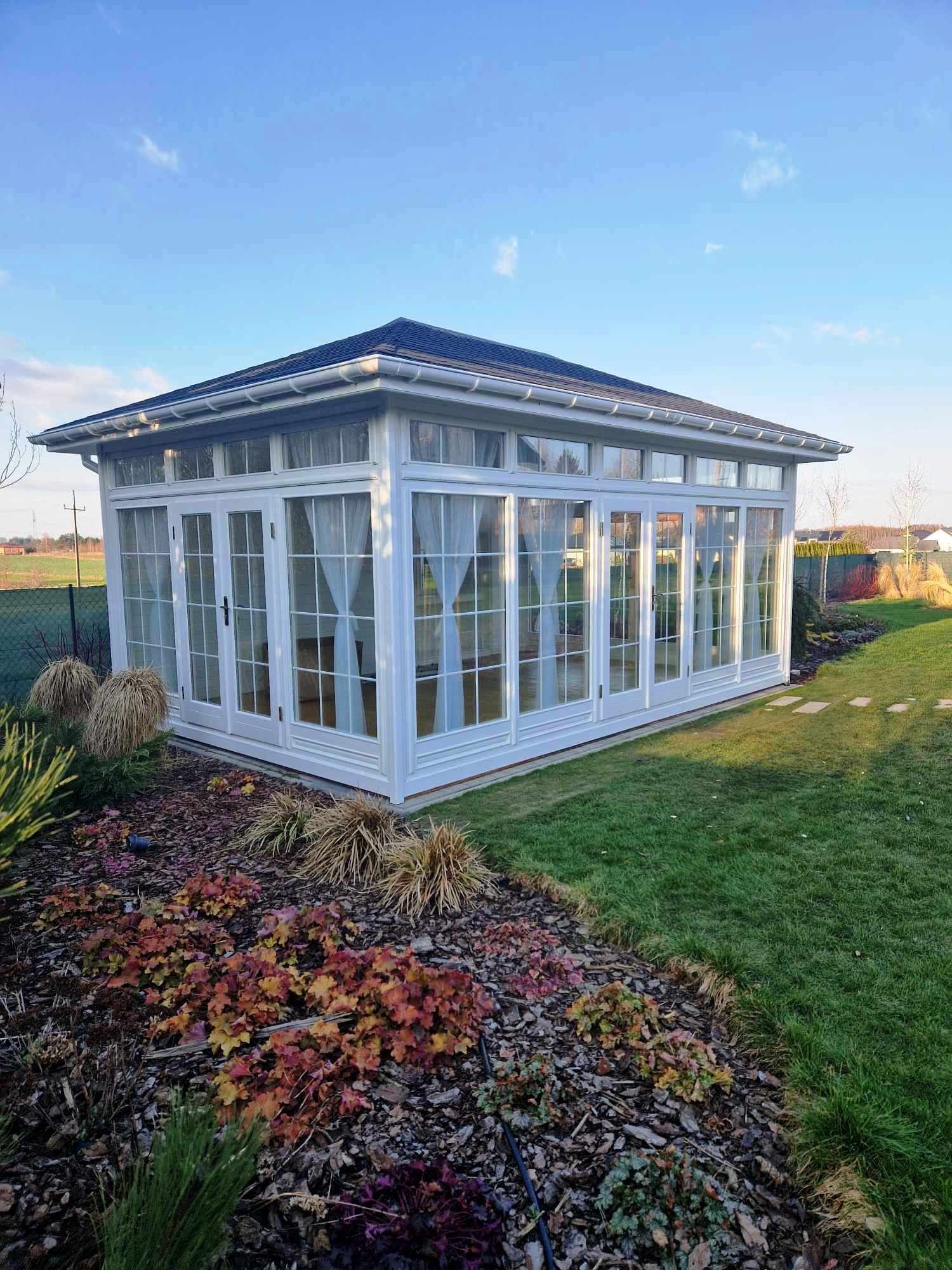 Biała, przeszklona konstrukcja z zasłonami wewnątrz, umieszczona w ogrodzie z rabatami i trawnikiem, pod błękitnym niebem.