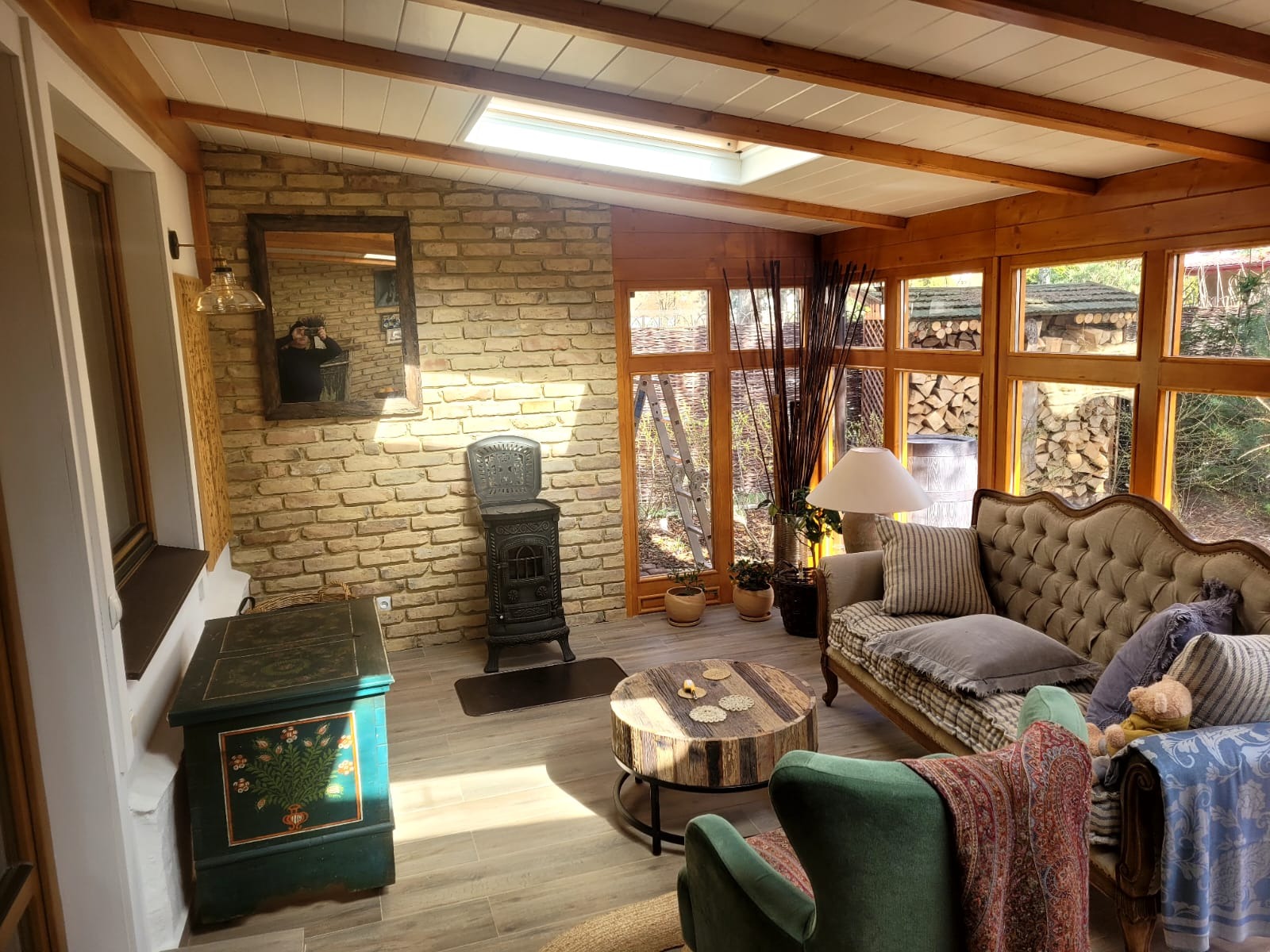 Wnętrze przeszklonej altany z ceglaną ścianą, piecem typu koza, drewnianym stropem i meblami w stylu vintage, widok na ogród z ułożonym drewnem.