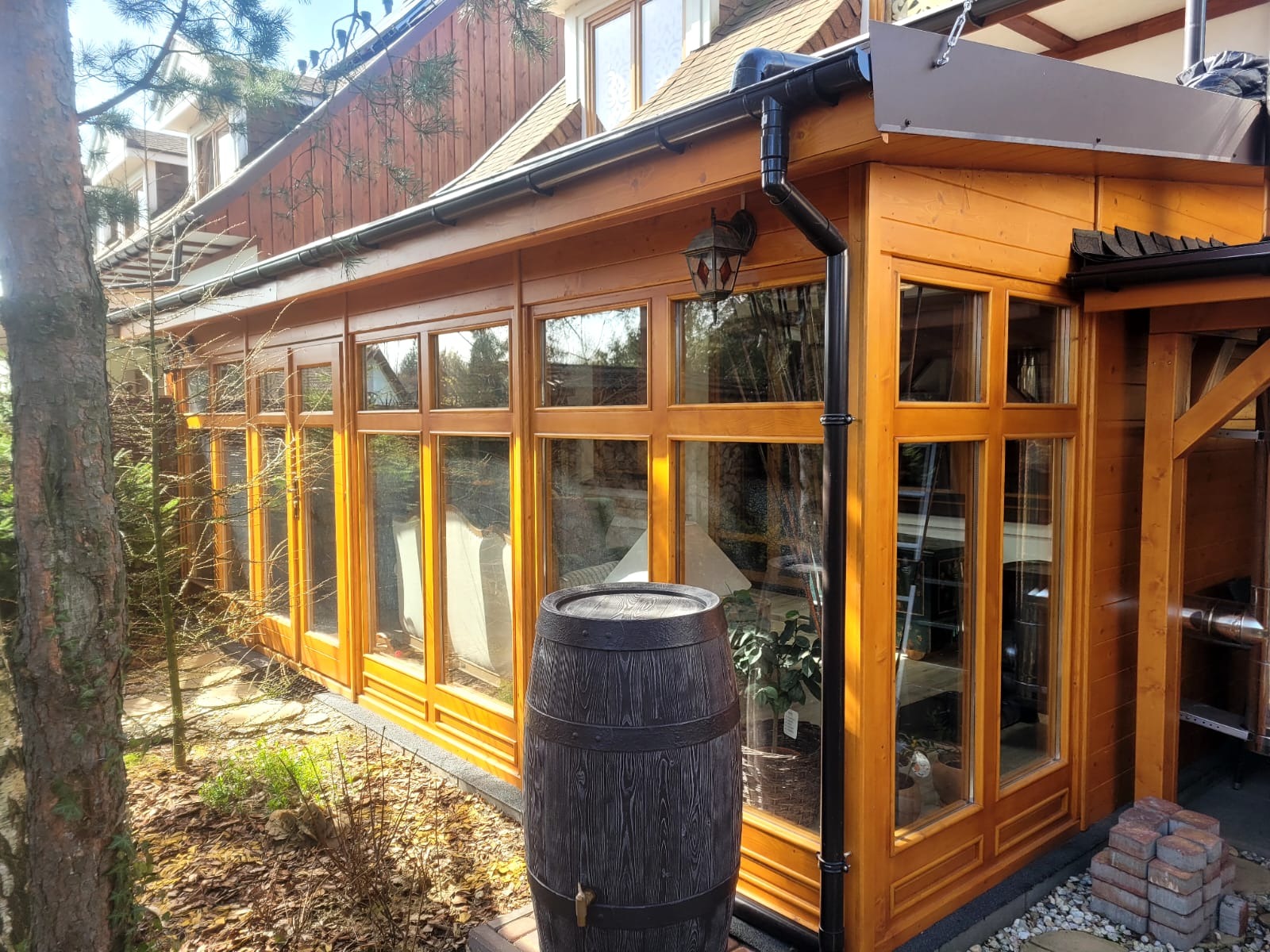 Drewniana konstrukcja oranżerii z licznymi oknami, widoczna z zewnątrz, z dekoracyjną beczką na pierwszym planie i fragmentem ogrodu.