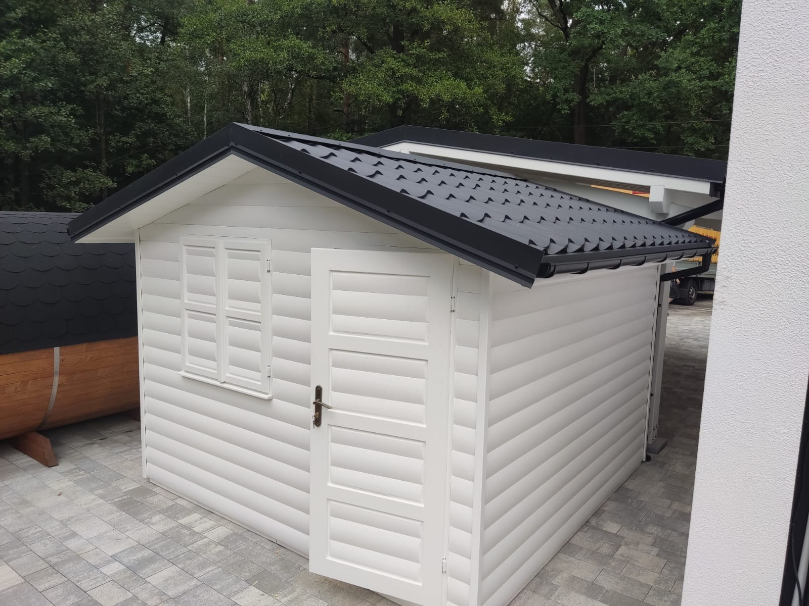 Biała altana ogrodowa z czarnym dachem, widok z boku, z widoczną drewnianą balią w tle.