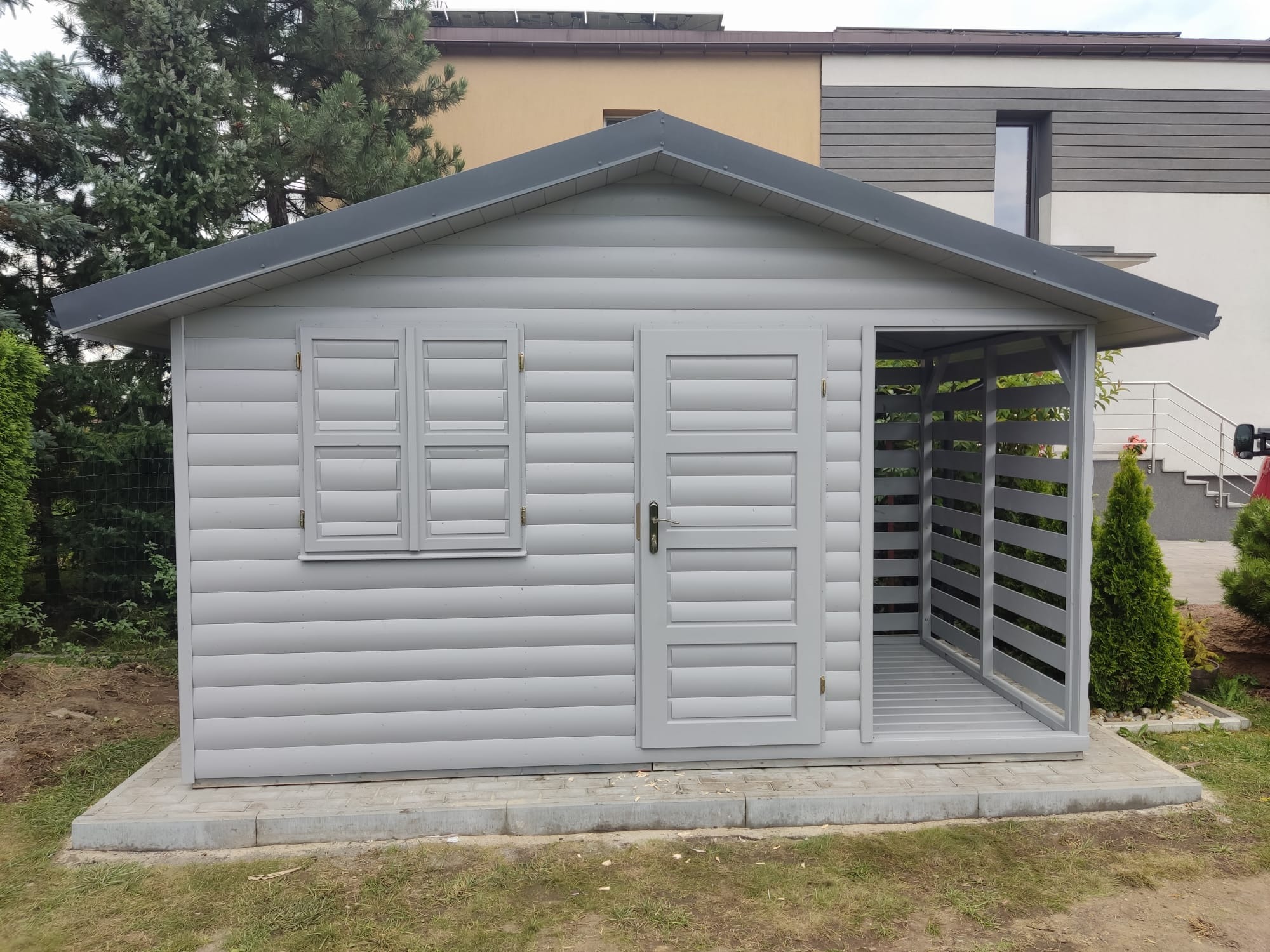 Szara, drewniana konstrukcja z oknem i otwartym wejściem, przypominająca mały domek ogrodowy, z widocznymi poziomymi deskami i ciemnym dachem, na tle zieleni i domu.