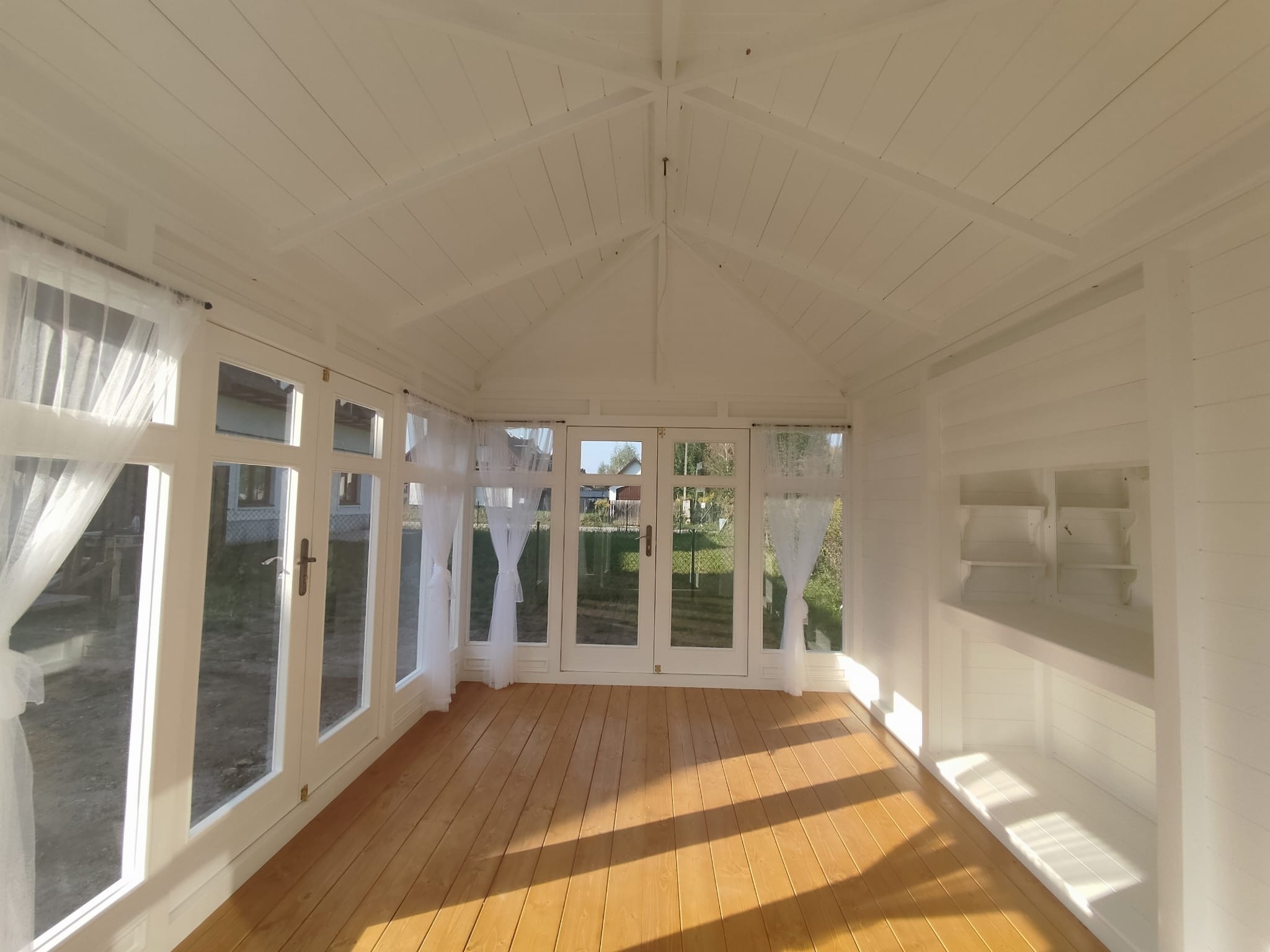 Jasne wnętrze altany z drewnianą podłogą, białymi ścianami i oknami z firankami, wpadające promienie słoneczne.