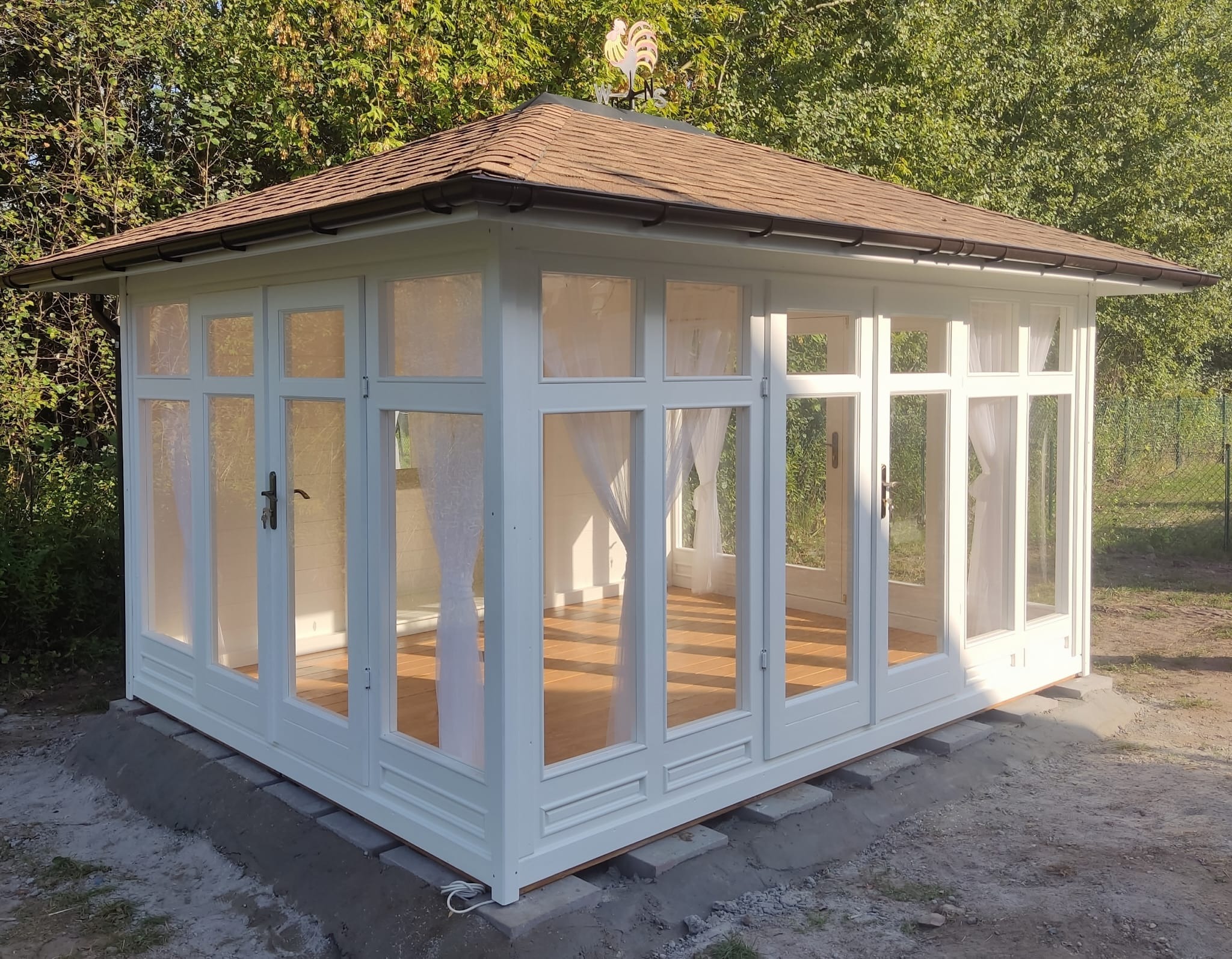 Biała, przeszklona altana z brązowym dachem i metalowym wiatrowskazem w kształcie koguta, umieszczona na betonowej podstawie w ogrodzie.