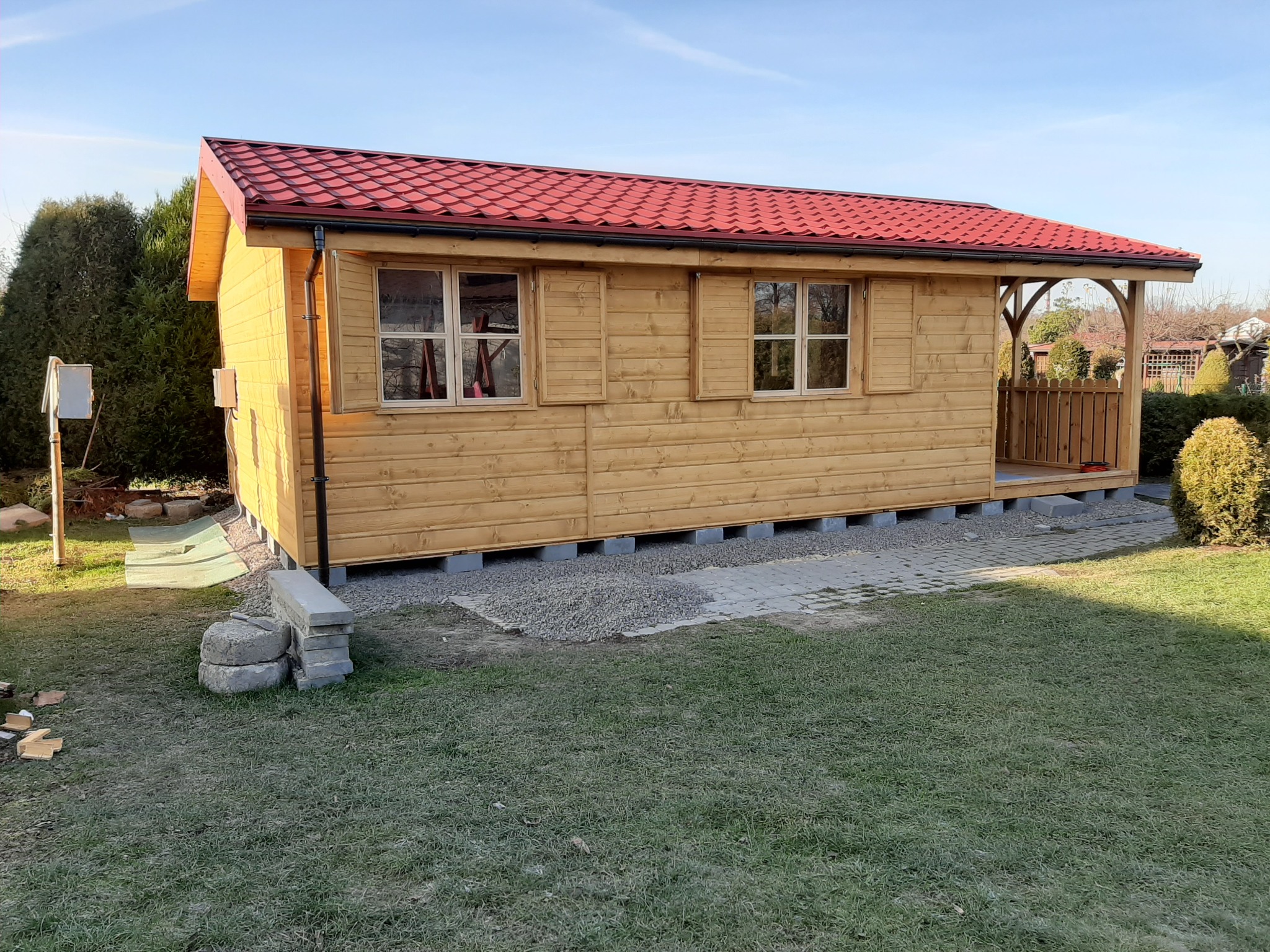 Świeżo postawiona, drewniana altana ogrodowa z czerwonym dachem w Skoczowie, osadzona na betonowych bloczkach, z otwartym wejściem i drewnianą balustradą.